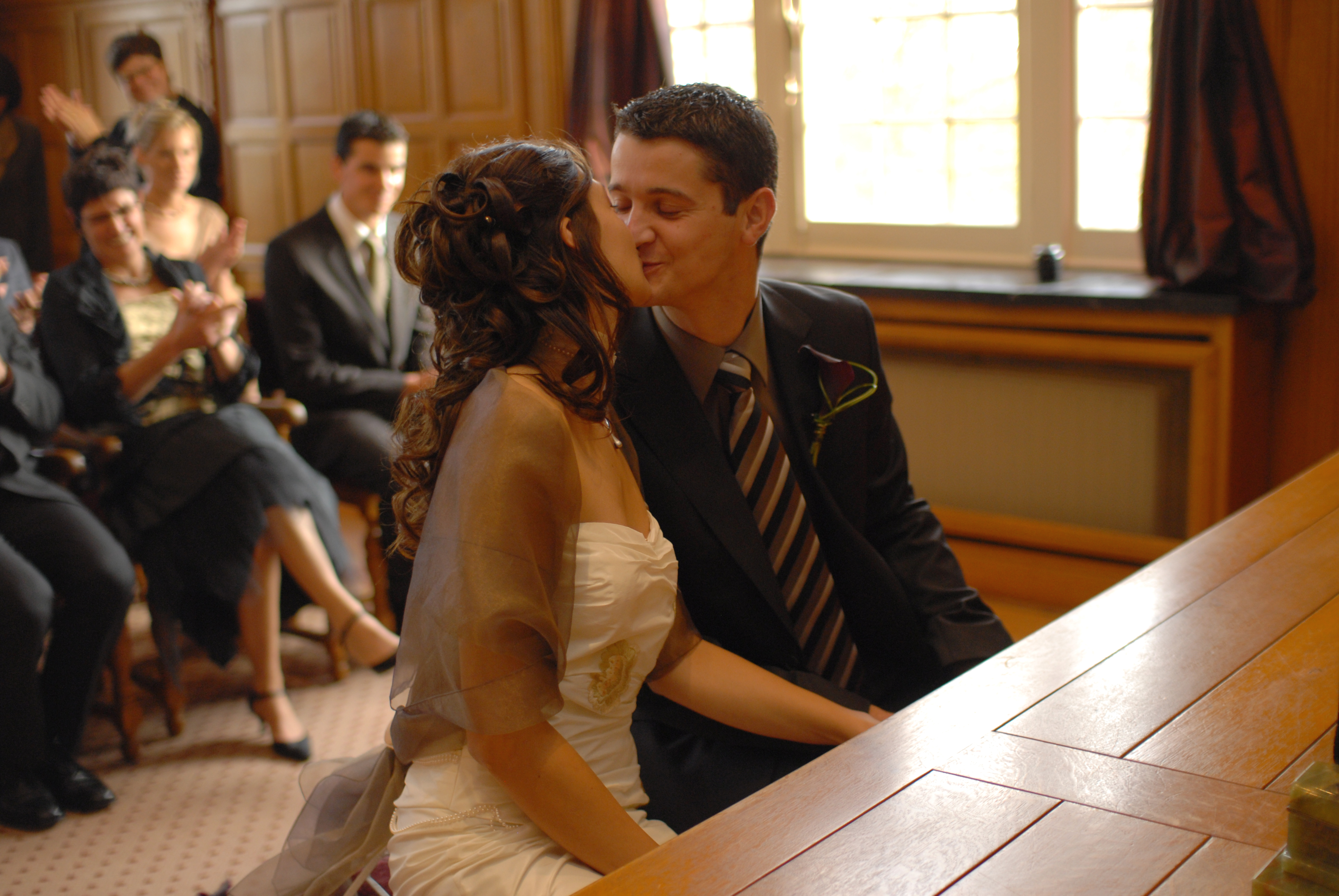 A bride and groom share a kiss during their wedding ceremony, while guests in the background clap and smile.