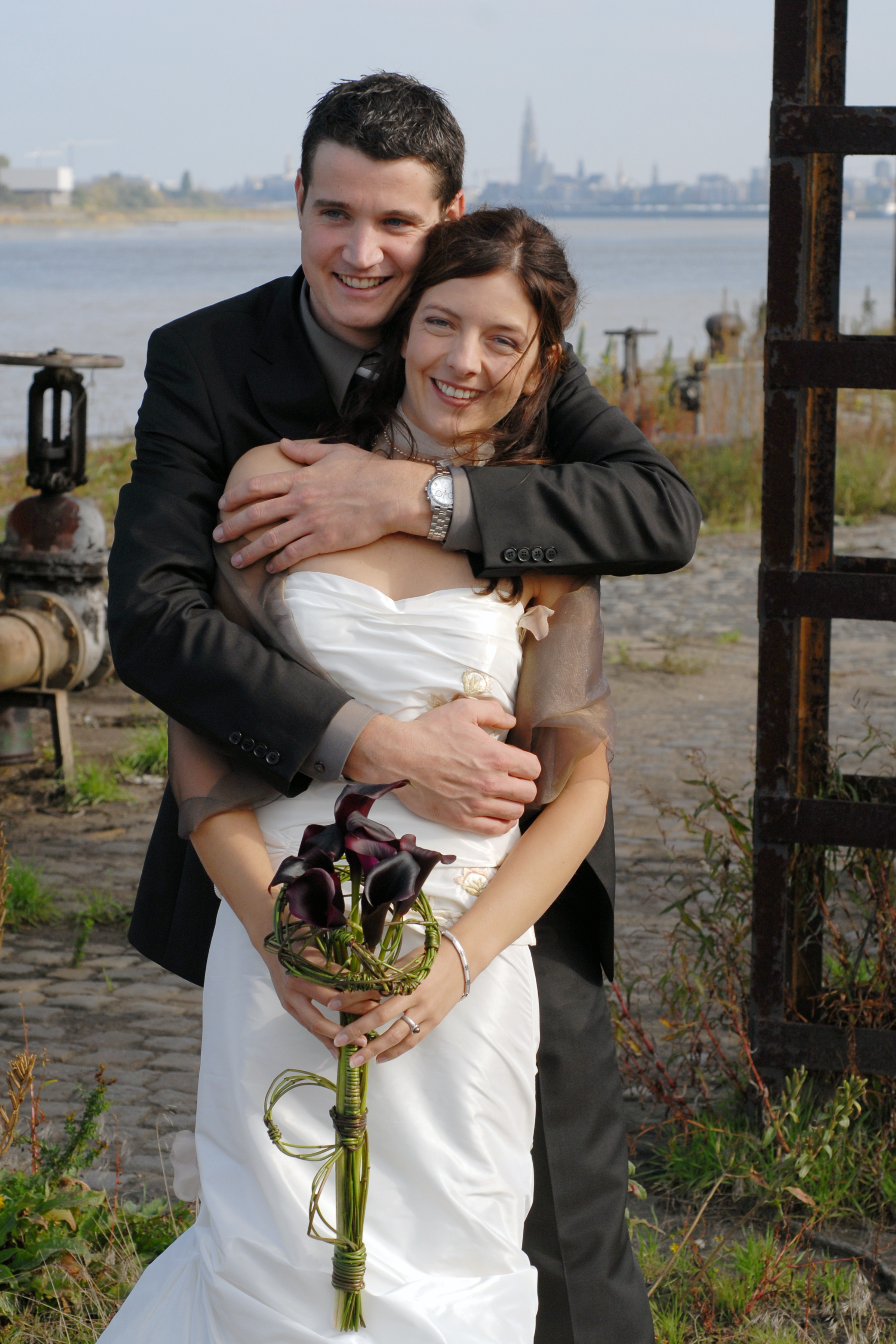 A smiling bride and groom embrace outdoors, with the bride holding a bouquet of dark flowers.