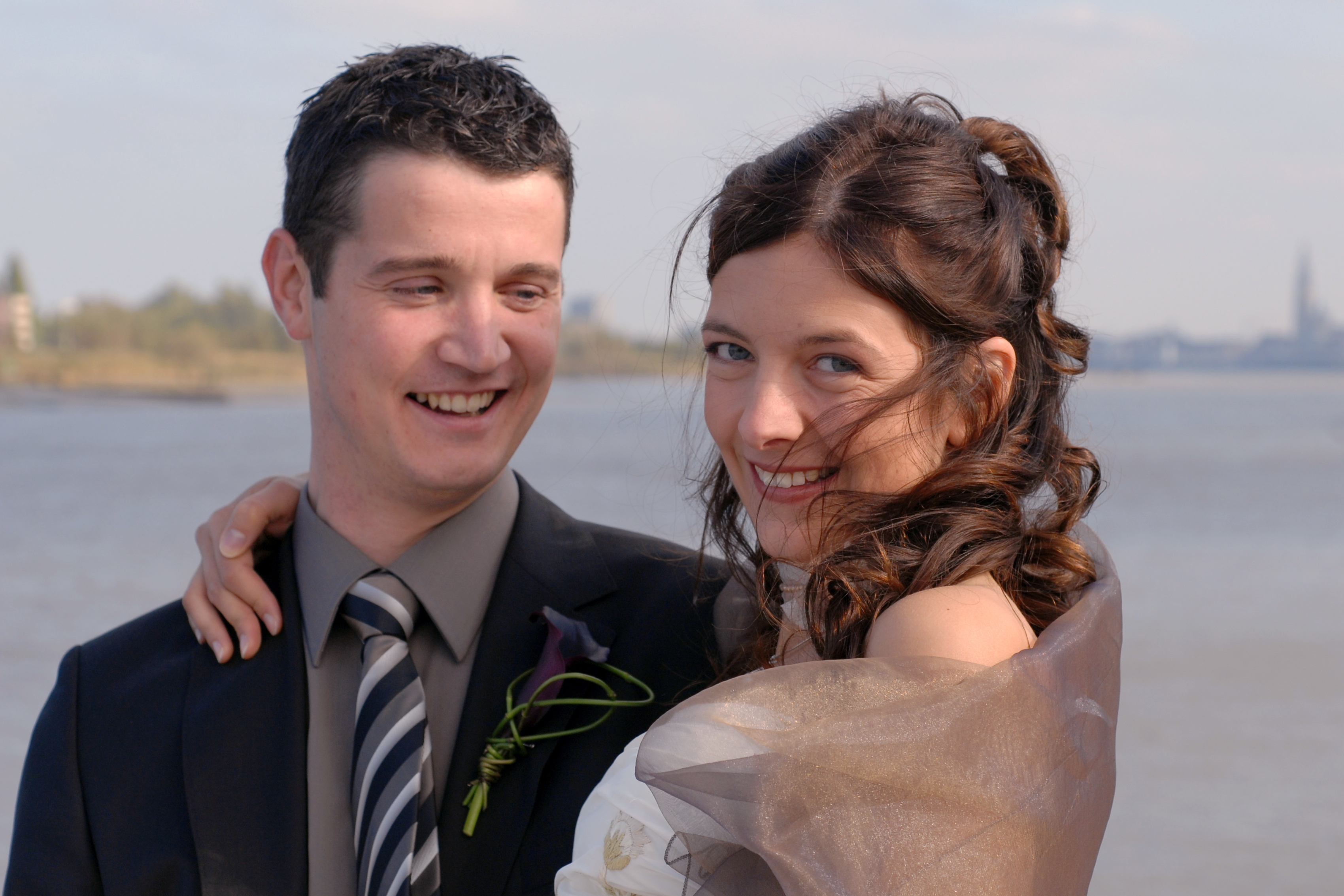 A smiling bride and groom pose together outdoors near the water on their wedding day.