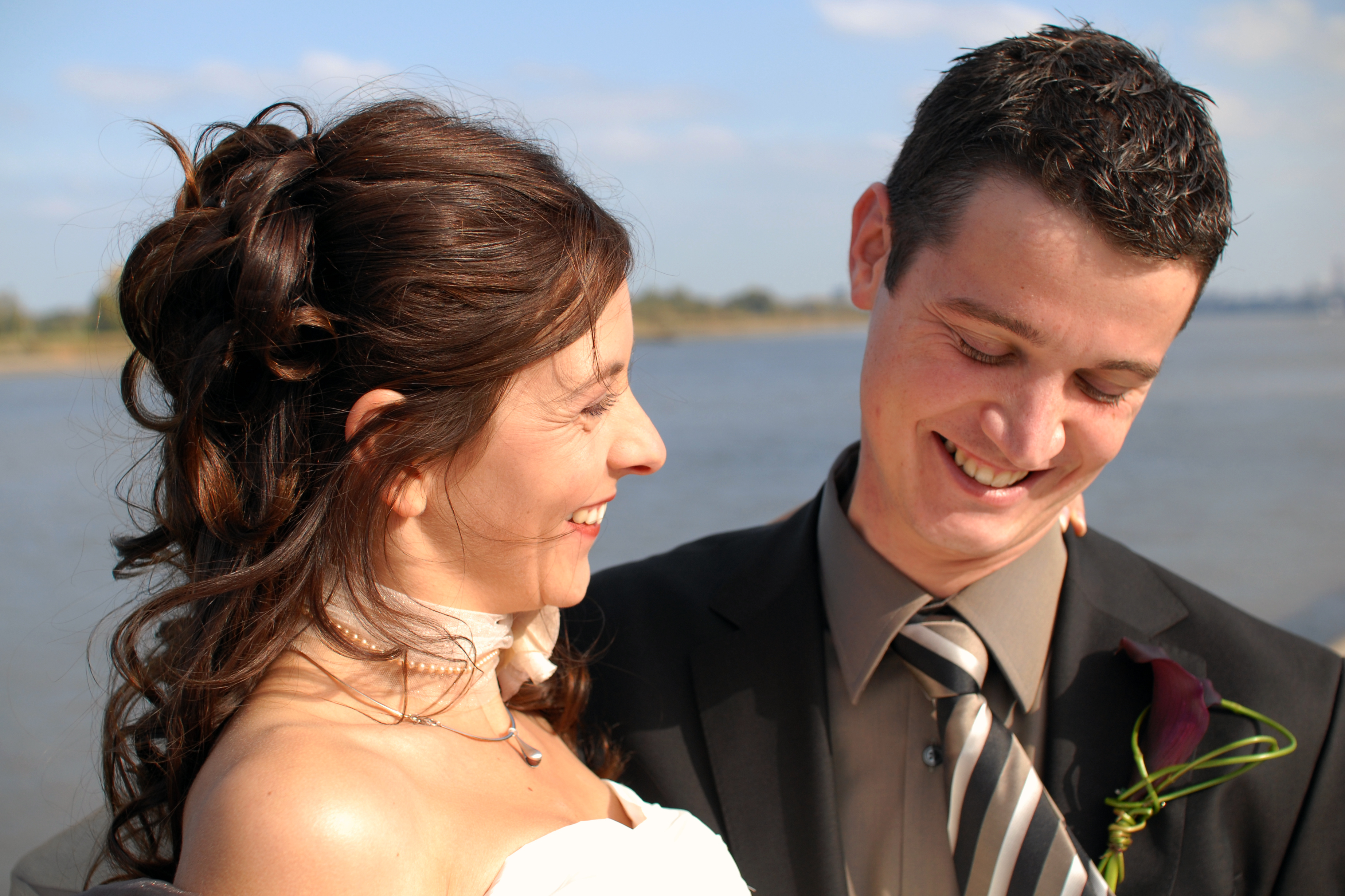 A bride and groom smile at each other outdoors, with the groom looking down and the bride leaning toward him.