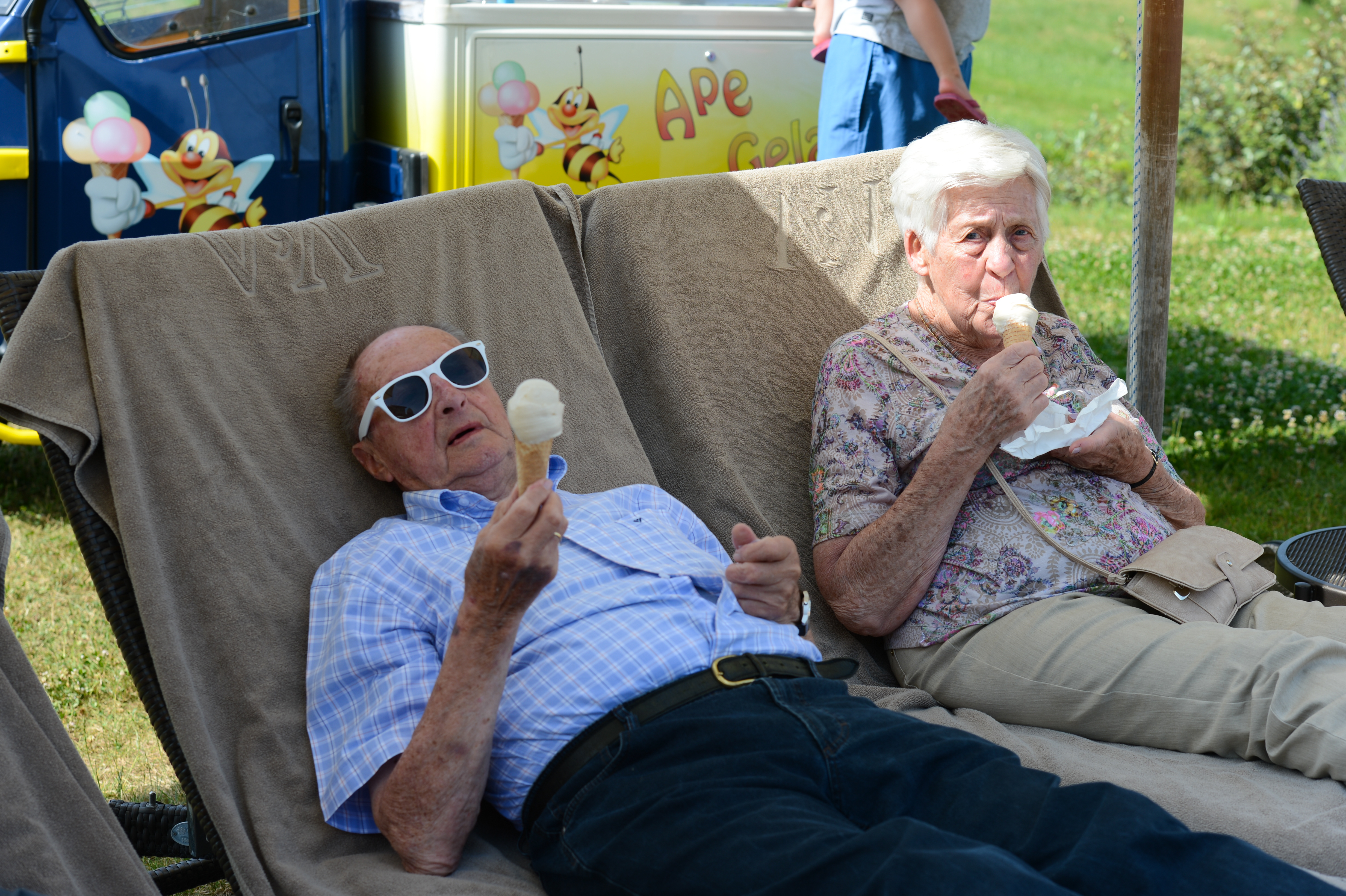 An elderly couple relaxes on lounge chairs, enjoying ice cream at an outdoor wedding celebration.