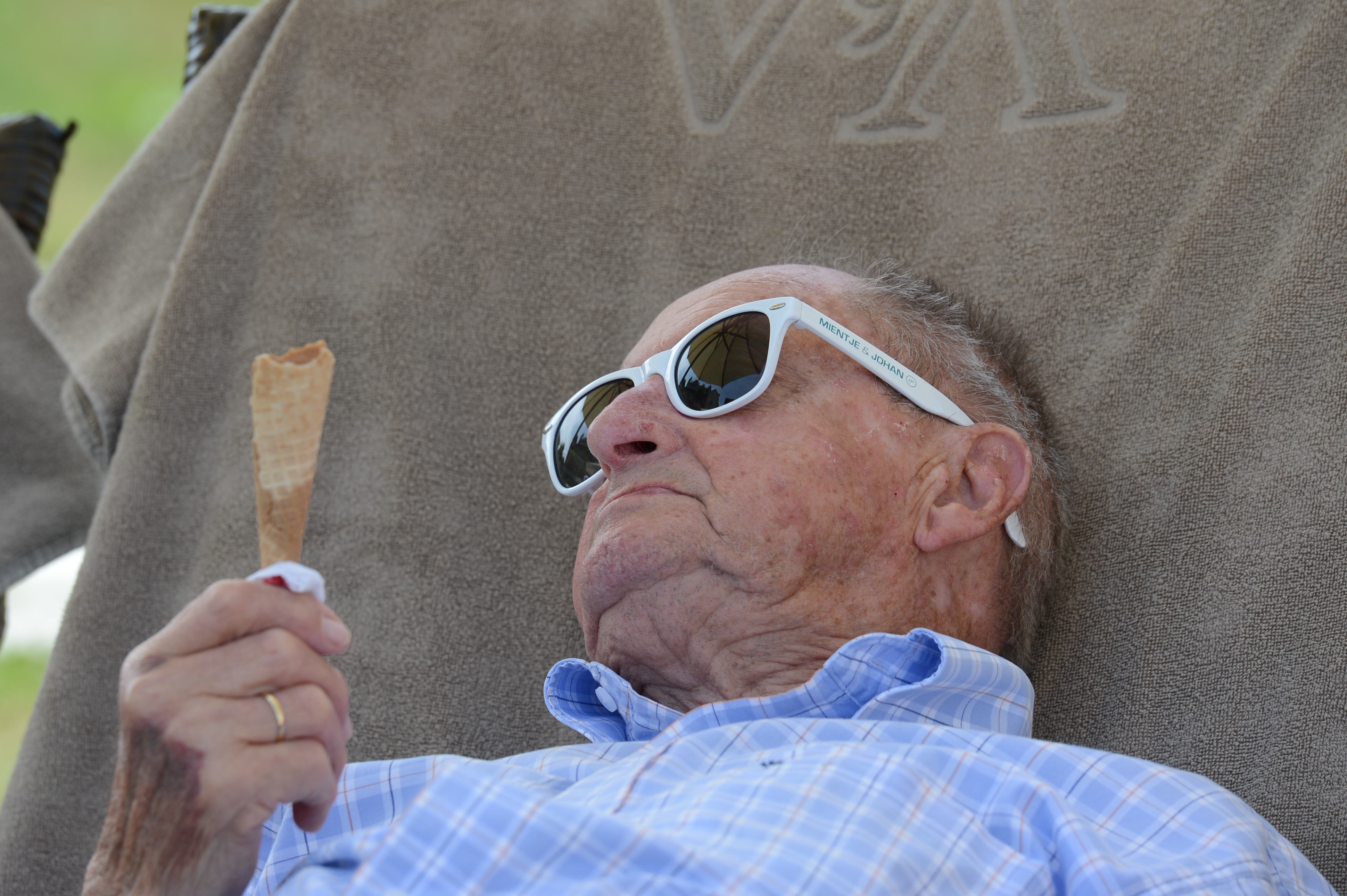 Elderly man wearing sunglasses reclines in a chair, holding an empty ice cream cone and looking at it.
