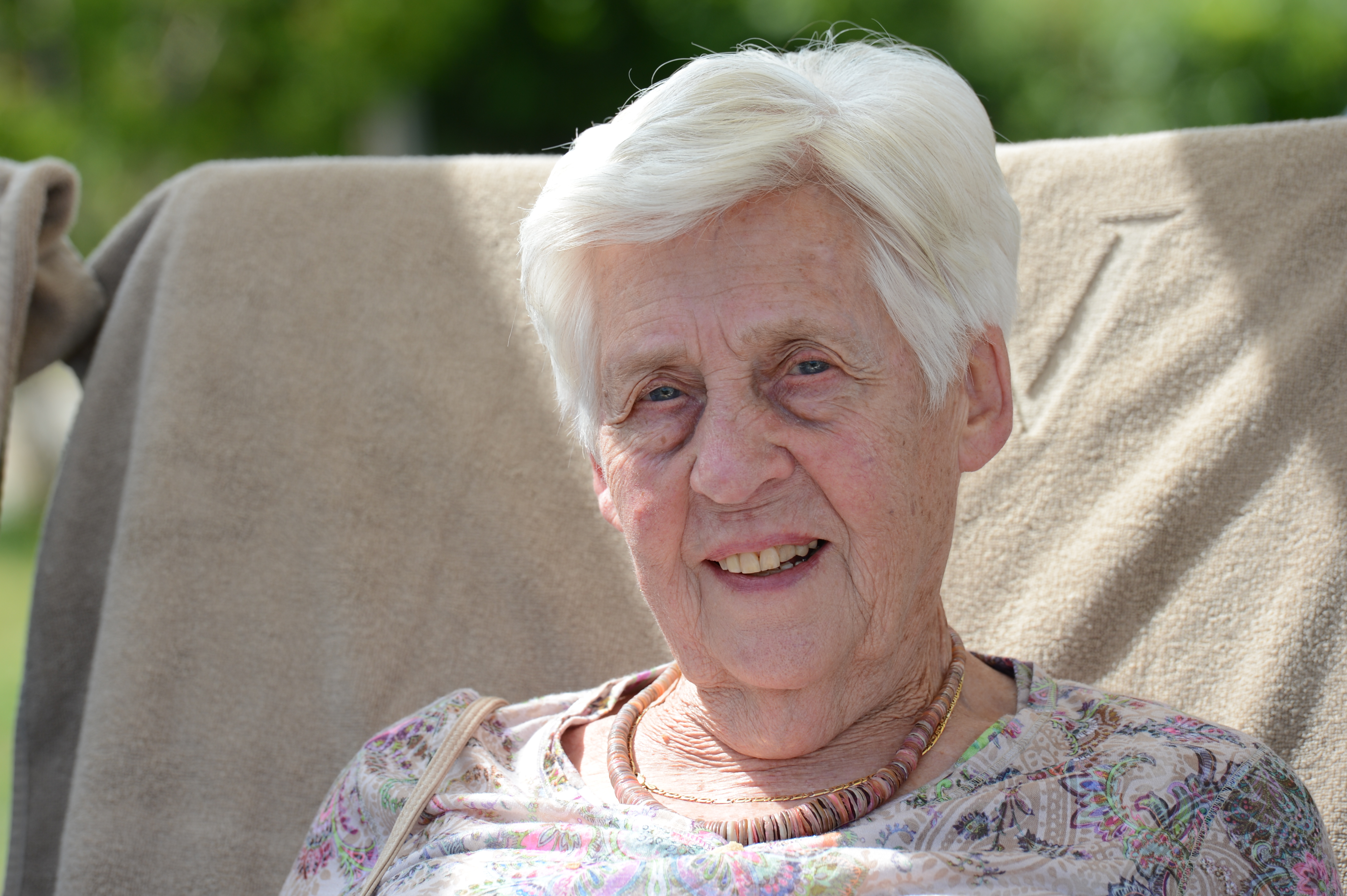 Elderly woman with white hair sits outdoors on a chair, smiling at the camera.