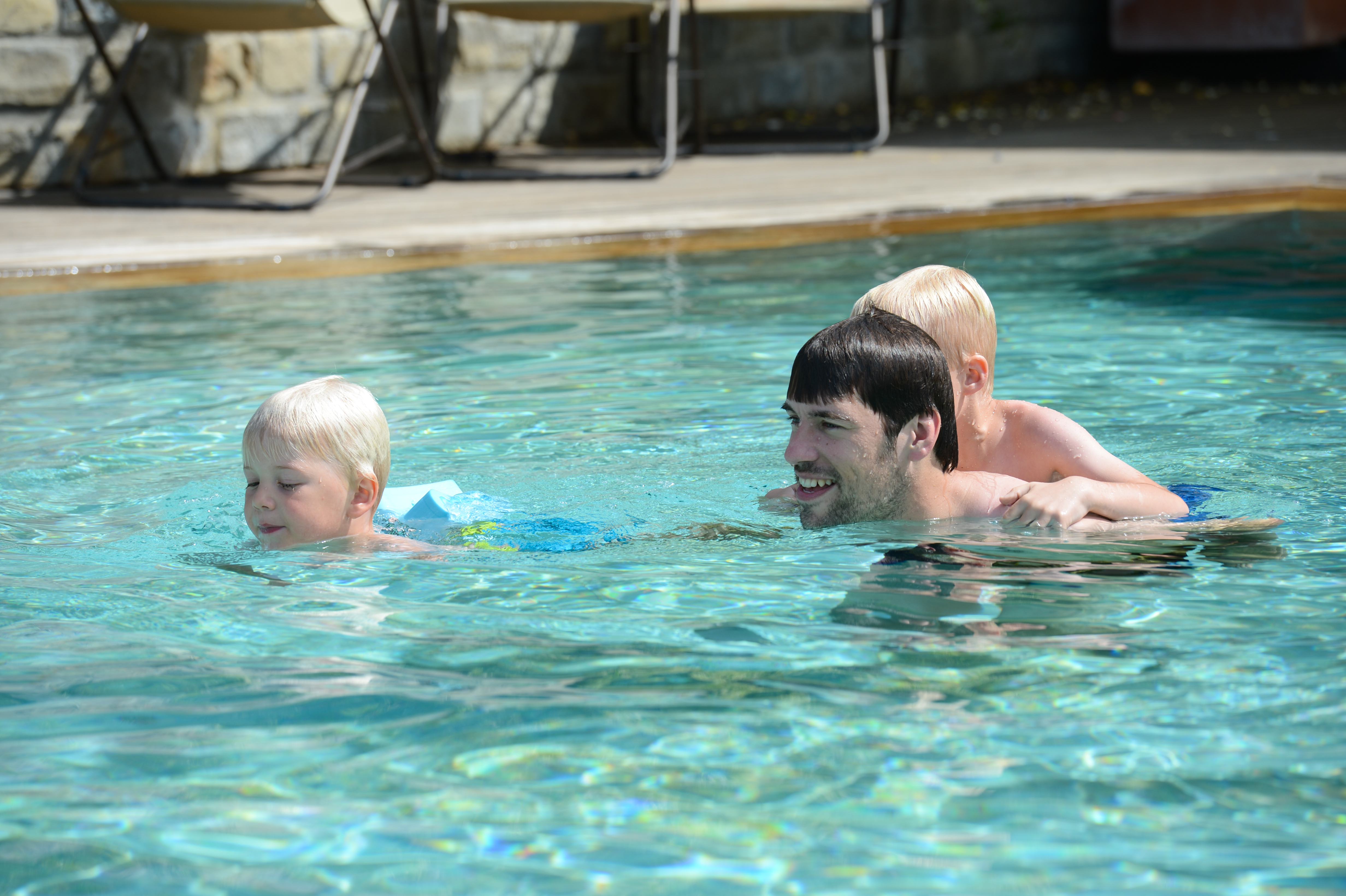 A man swims in a pool with two children, one holding onto his back while another swims ahead.