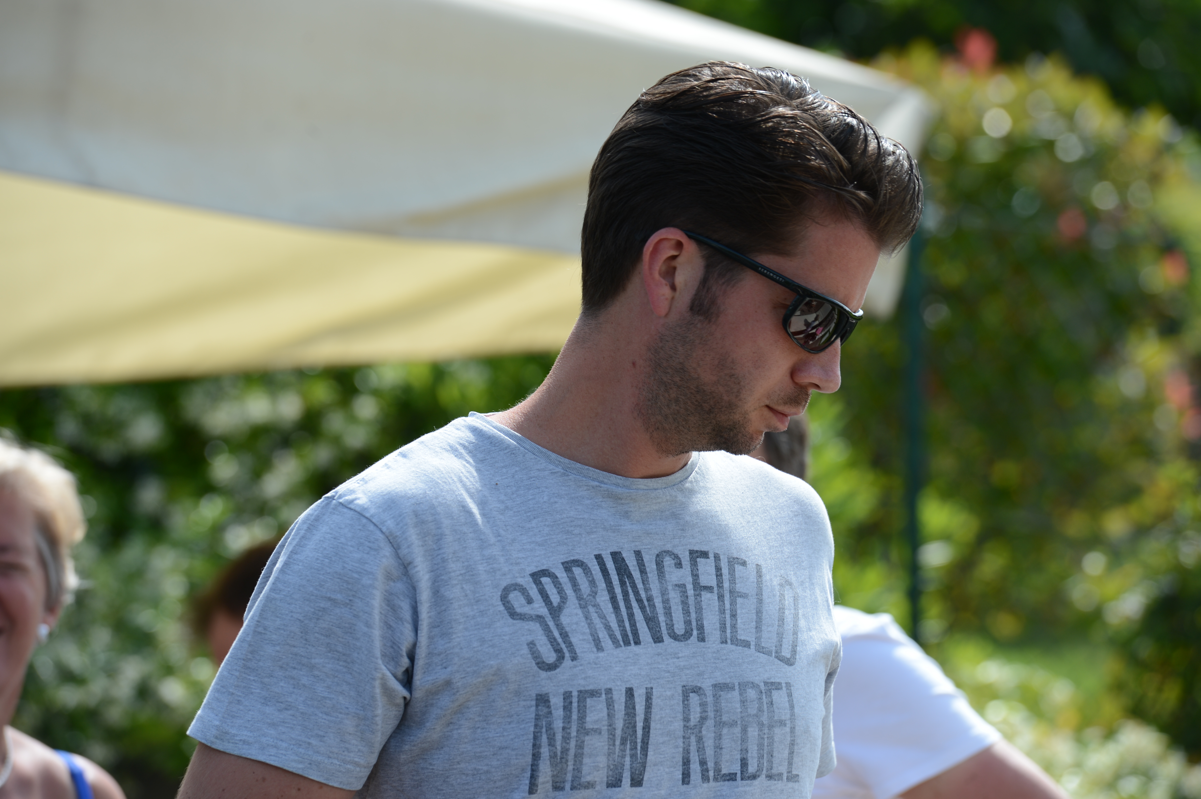 A man wearing sunglasses and a casual t-shirt looks down while standing outdoors at a wedding event.