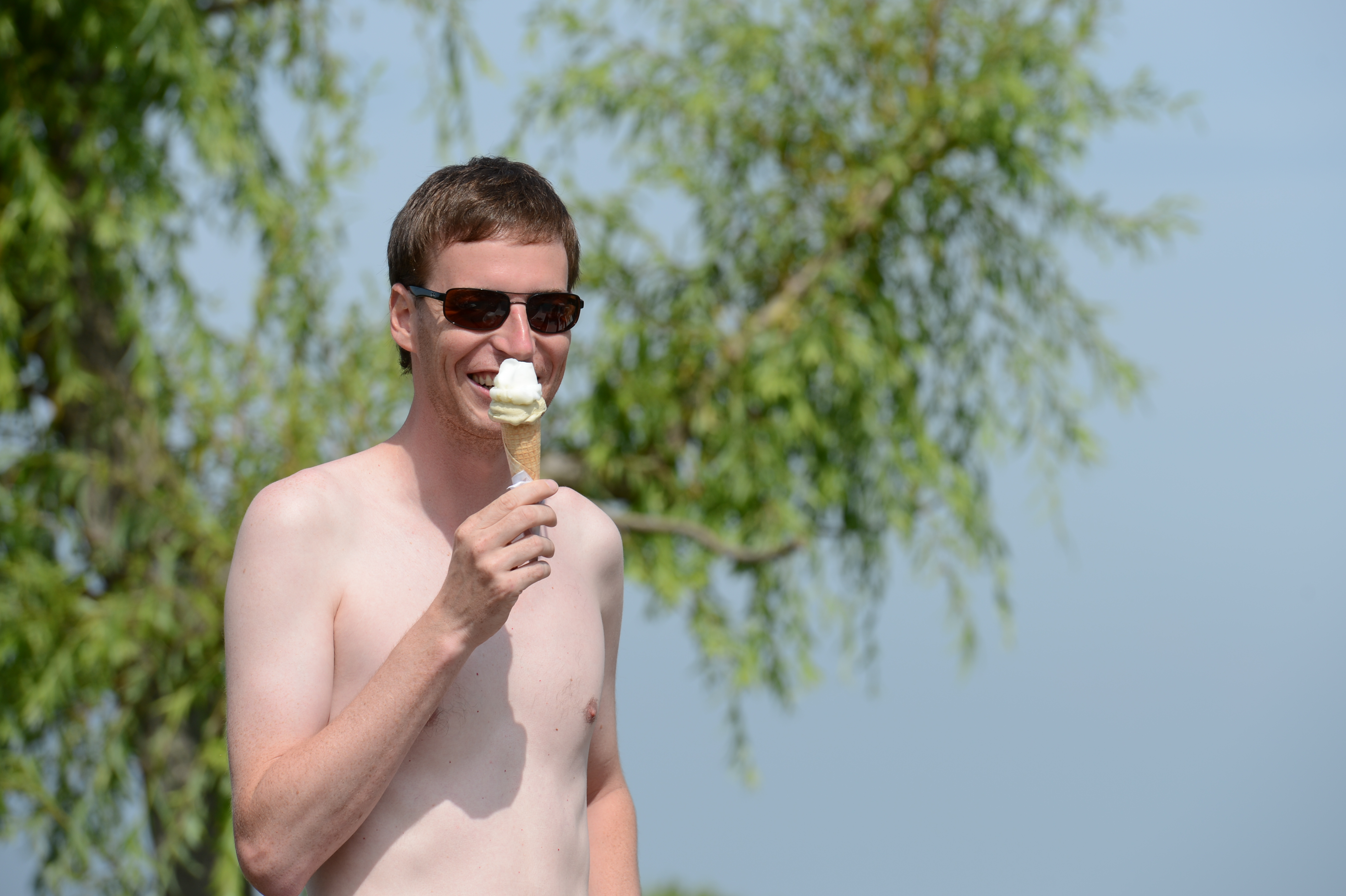 A shirtless man wearing sunglasses smiles while holding an ice cream cone outdoors.