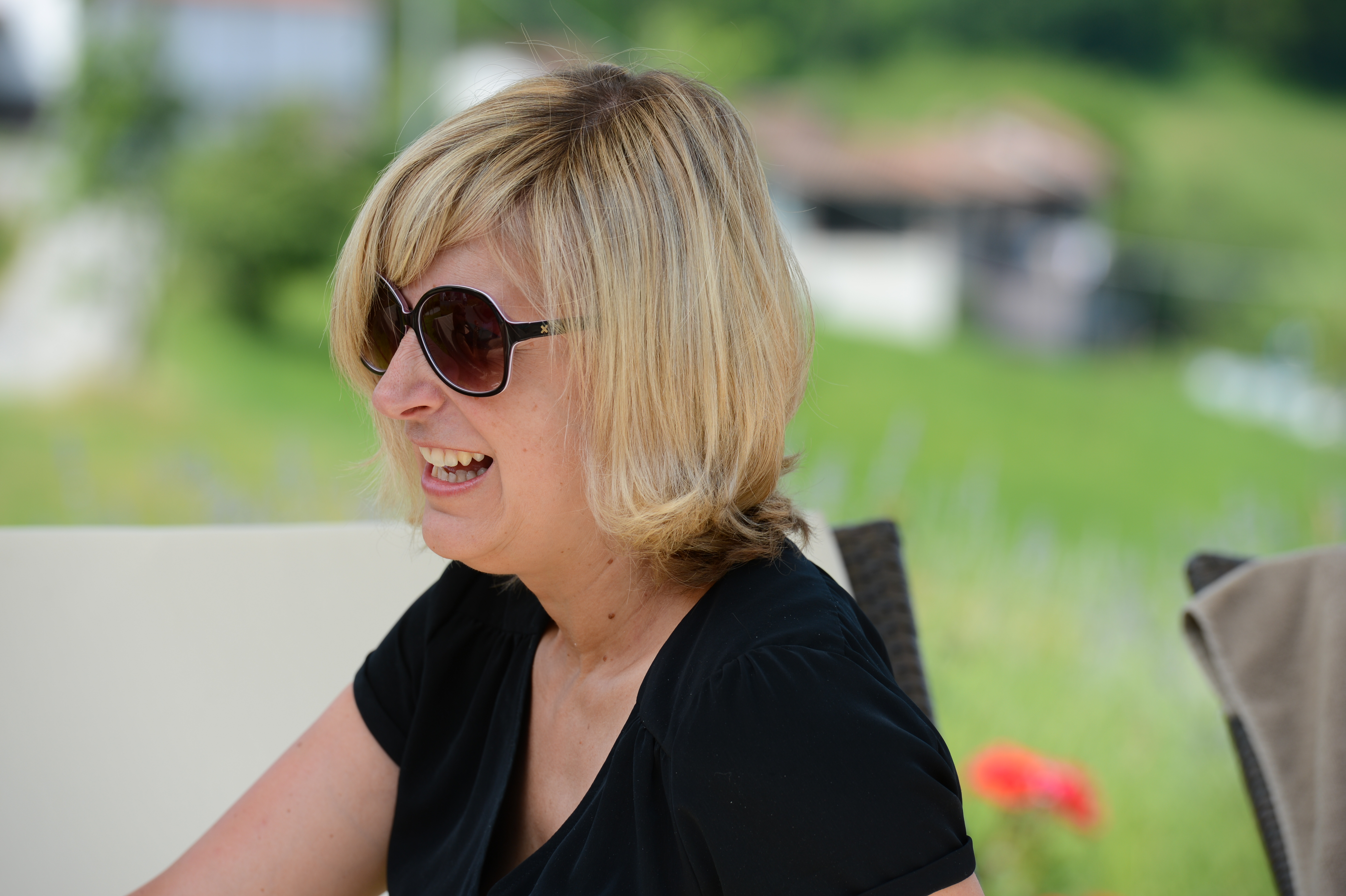 A woman wearing sunglasses and a black dress smiles while sitting outdoors at a wedding.