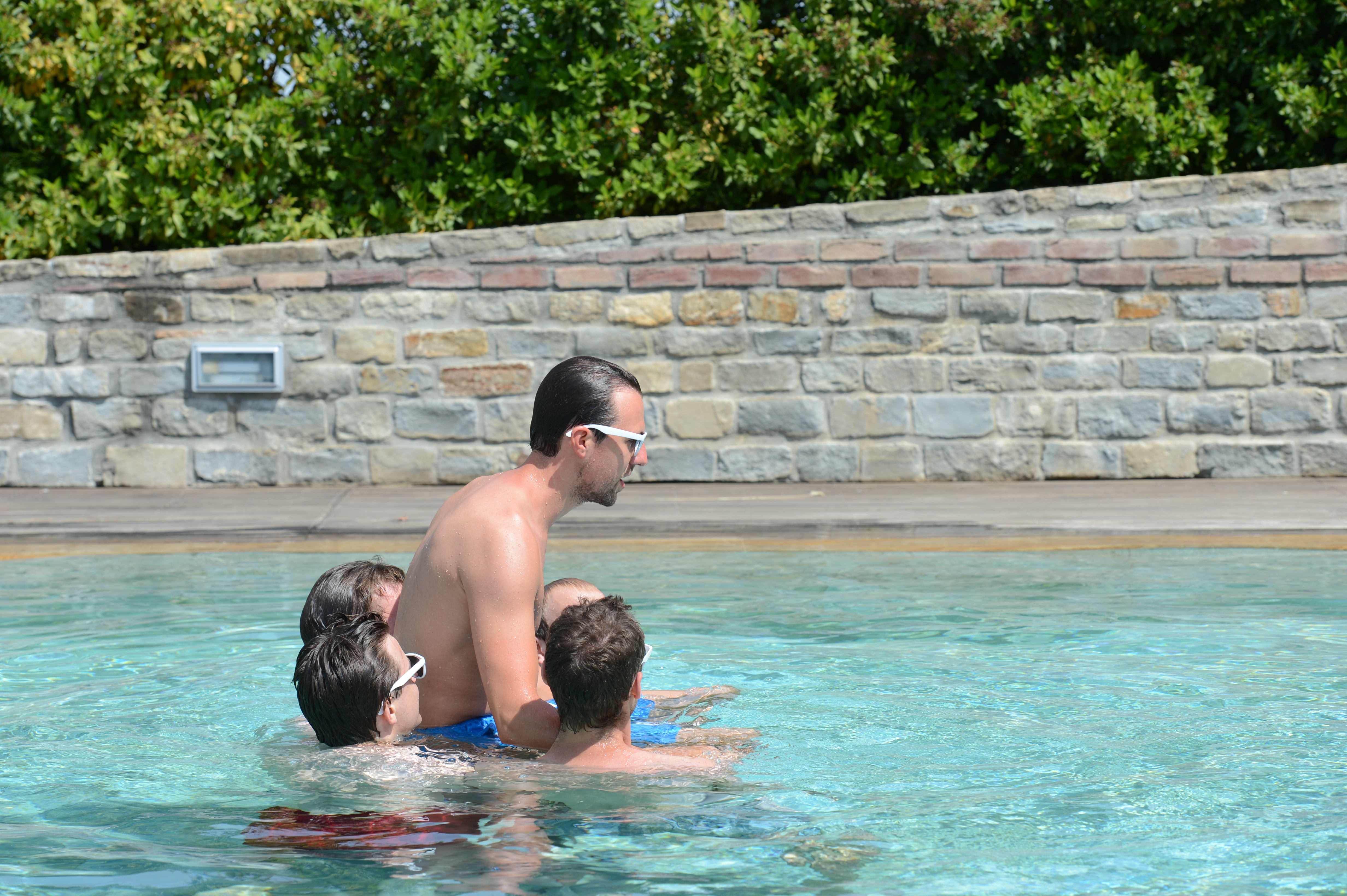 A group of people wearing sunglasses play together in a swimming pool during a wedding celebration.