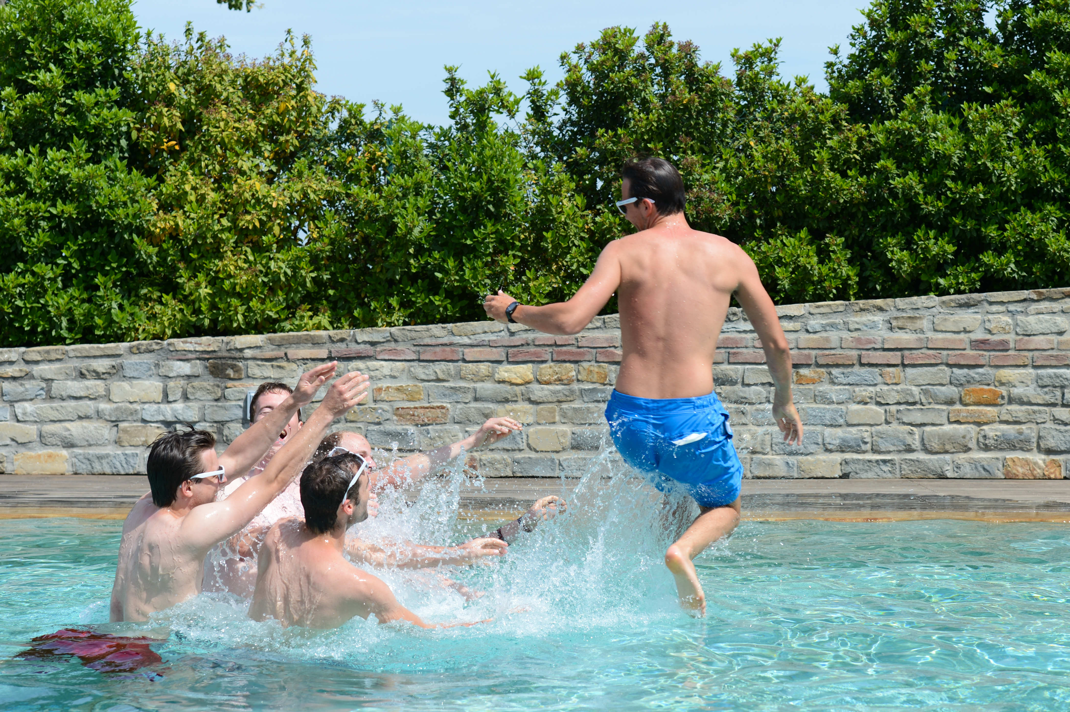 A man in blue shorts jumps into a pool where several men reach out and splash water.