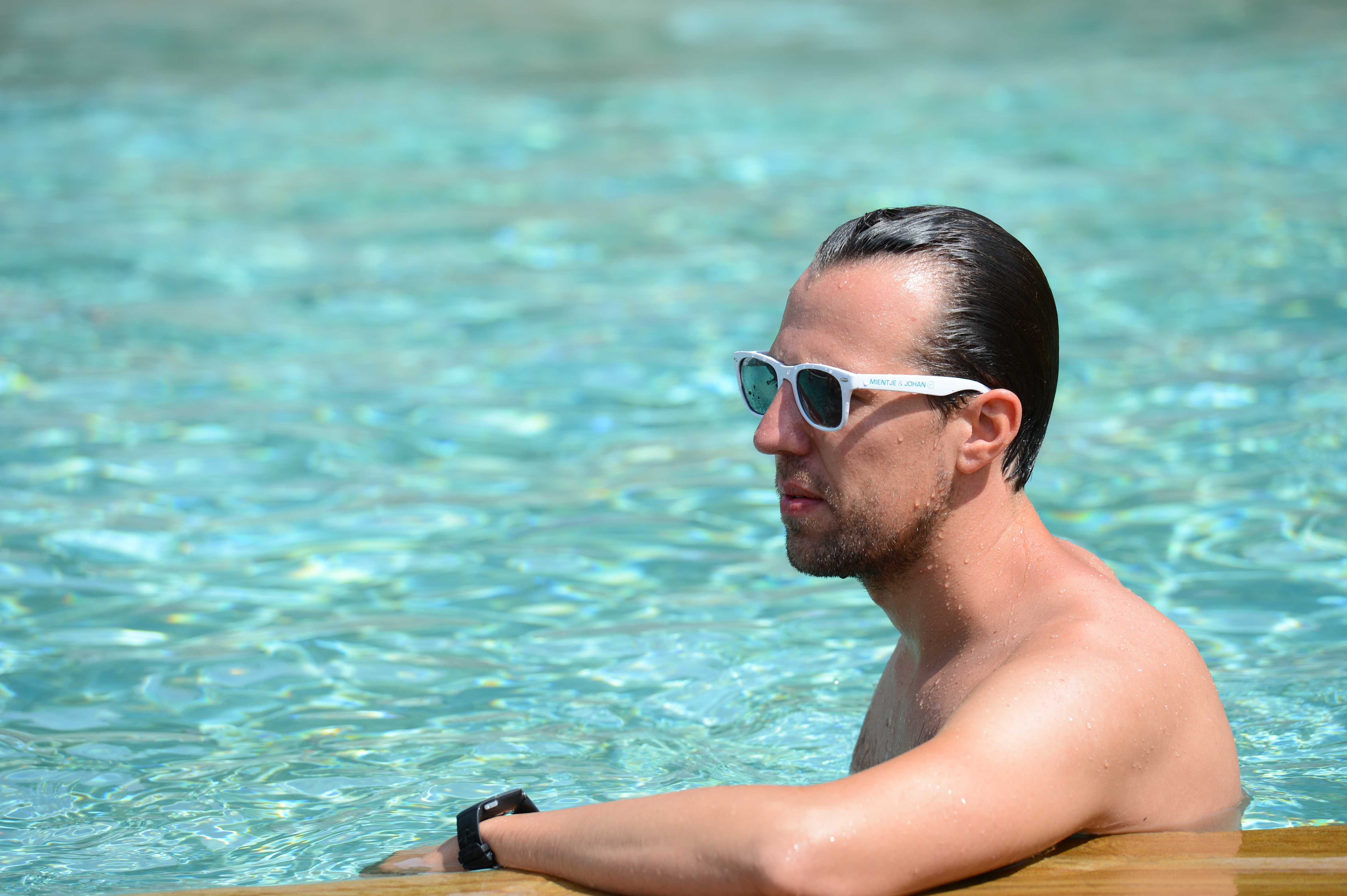 A man wearing sunglasses leans on the edge of a pool, partially submerged in the water.