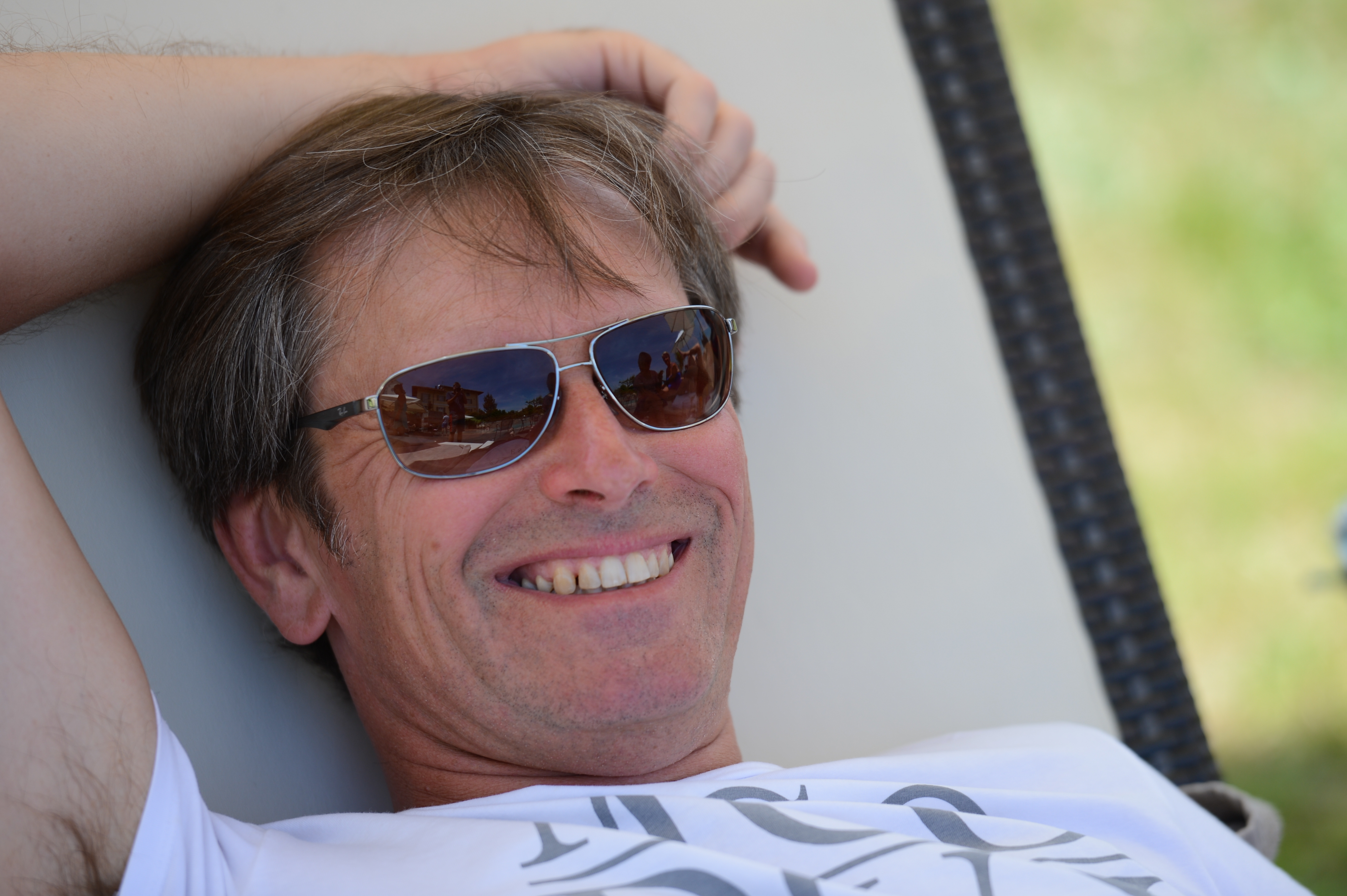 A man wearing sunglasses and a white shirt smiles while relaxing on a lounge chair outdoors.