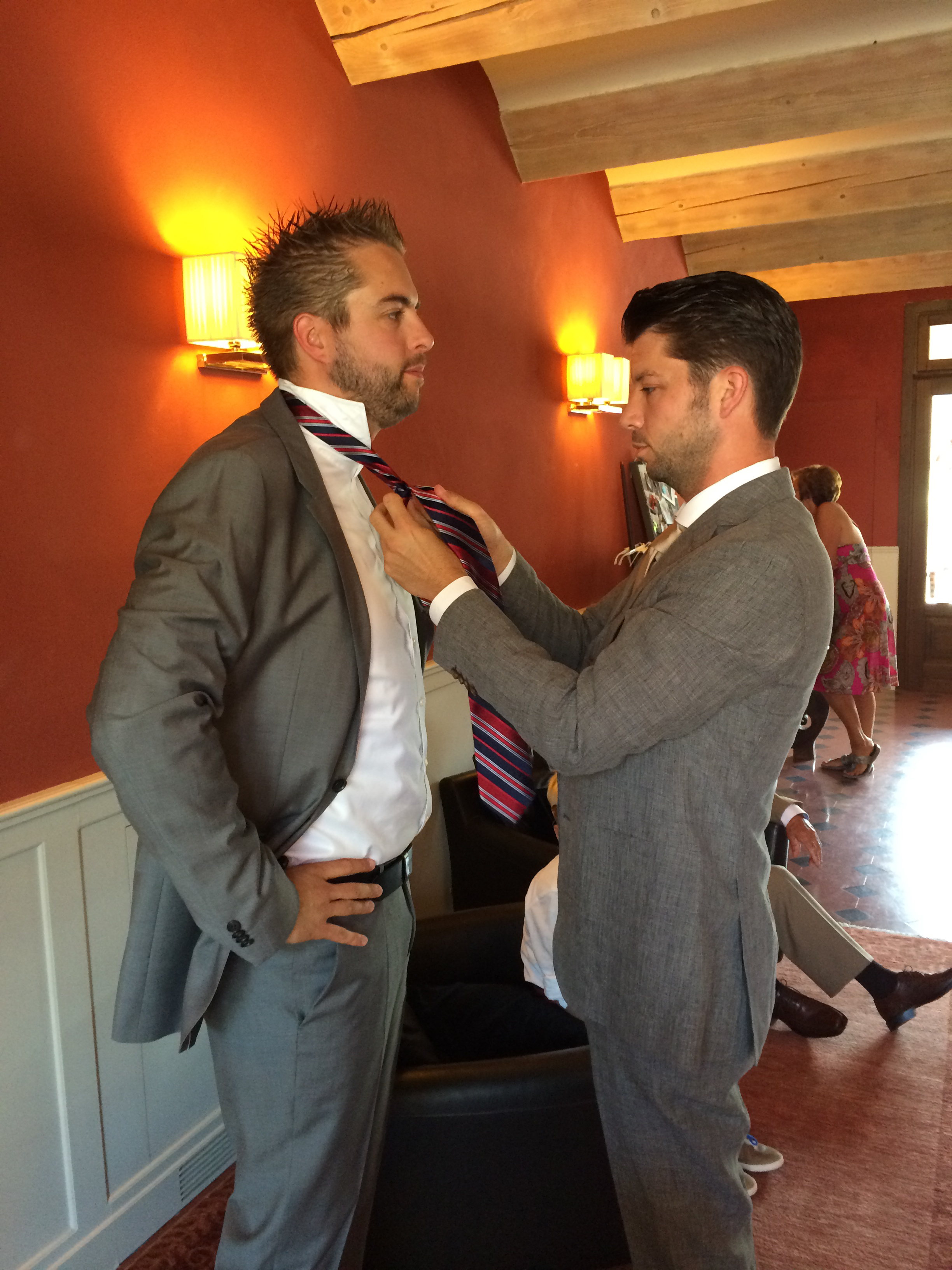 A man in a suit helps another man adjust his tie before a wedding.