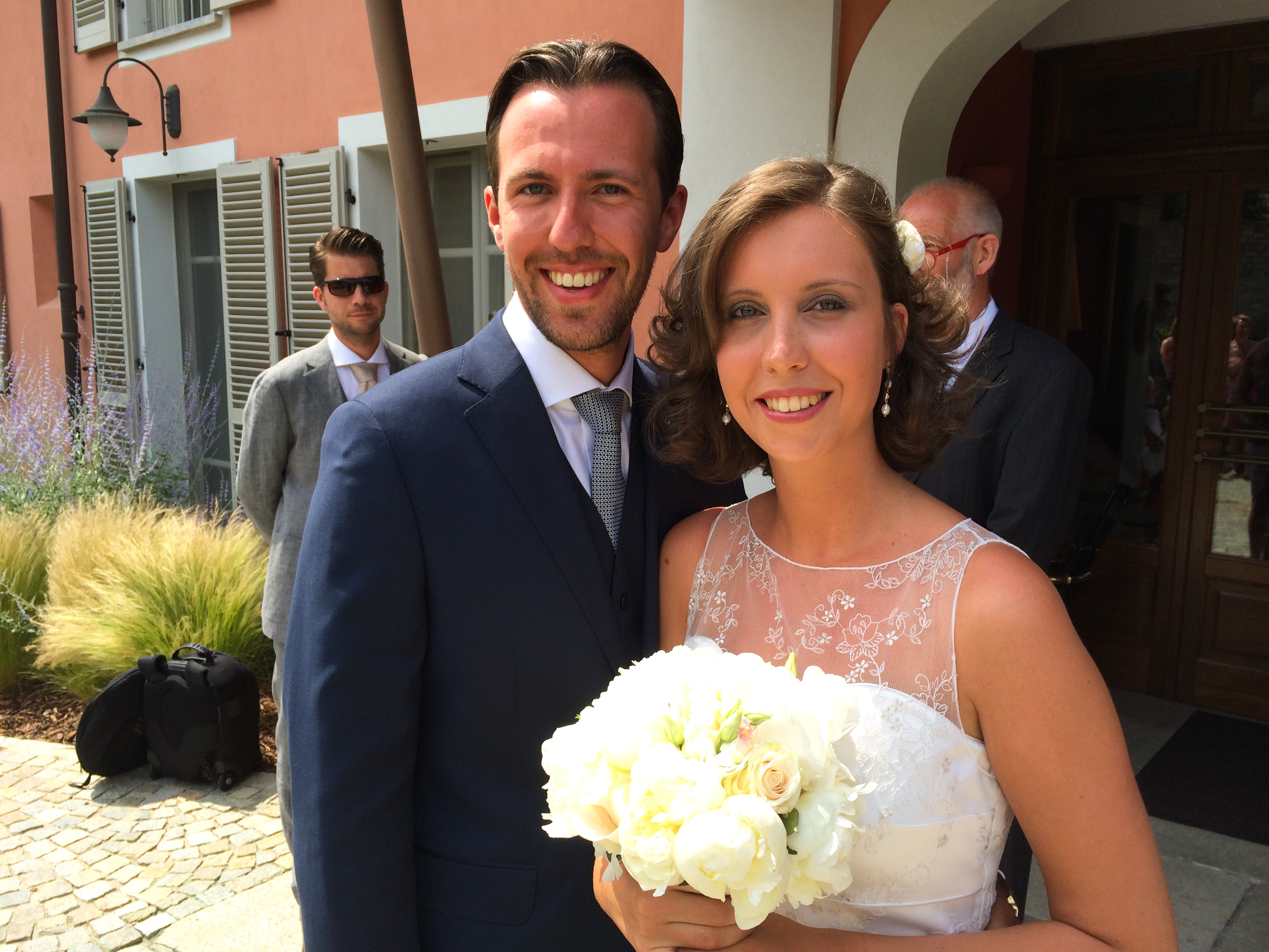A bride and groom smile at the camera, with the bride holding a bouquet of white flowers.