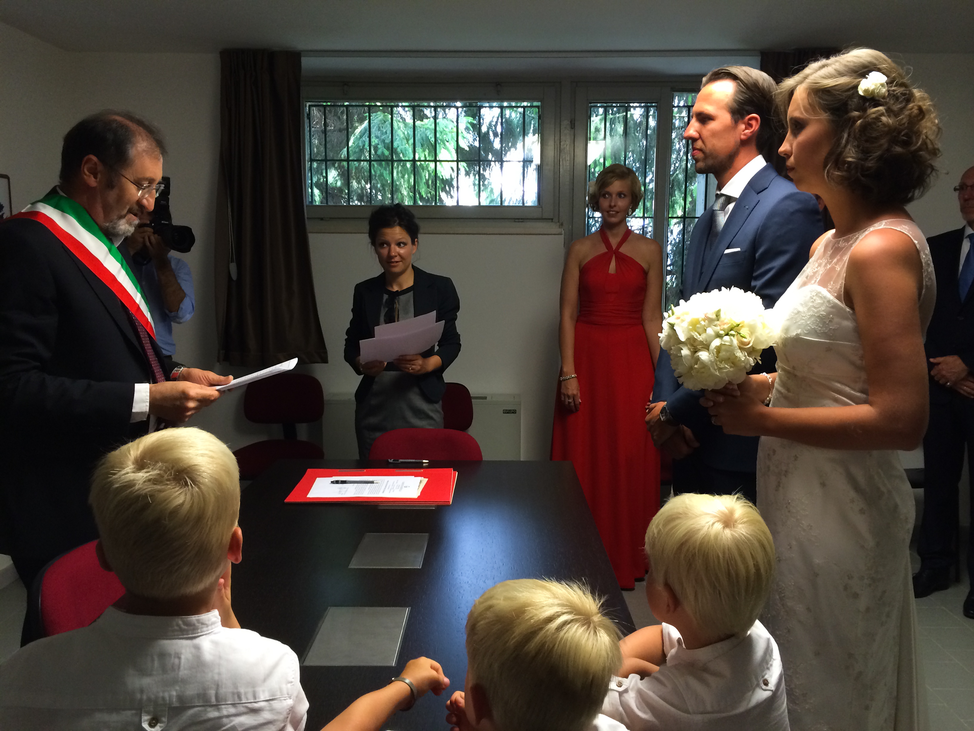 A bride and groom stand before an officiant during their wedding ceremony, holding flowers, with guests watching.