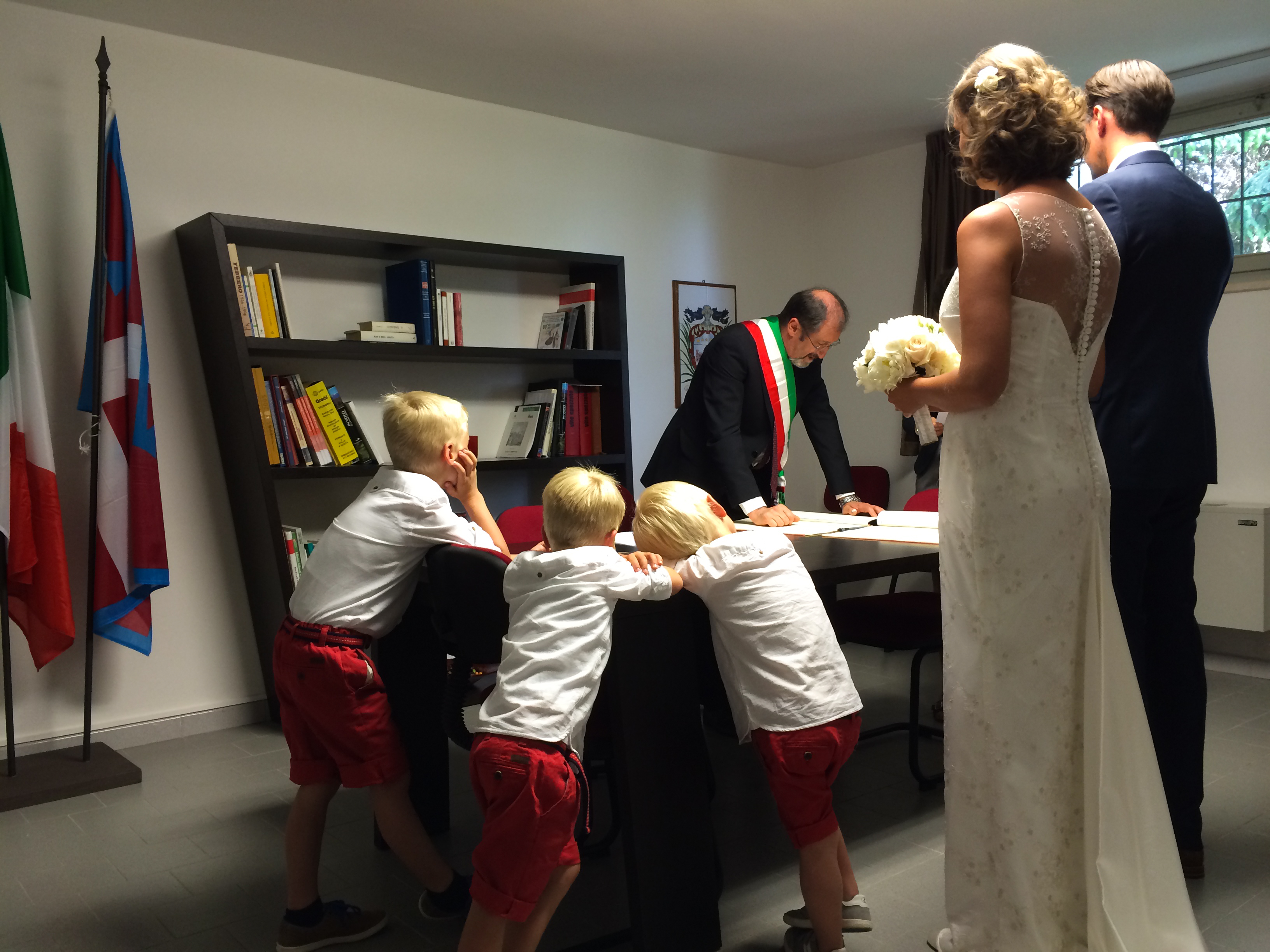 A bride and groom stand before an official signing documents, while three young boys watch from the side.