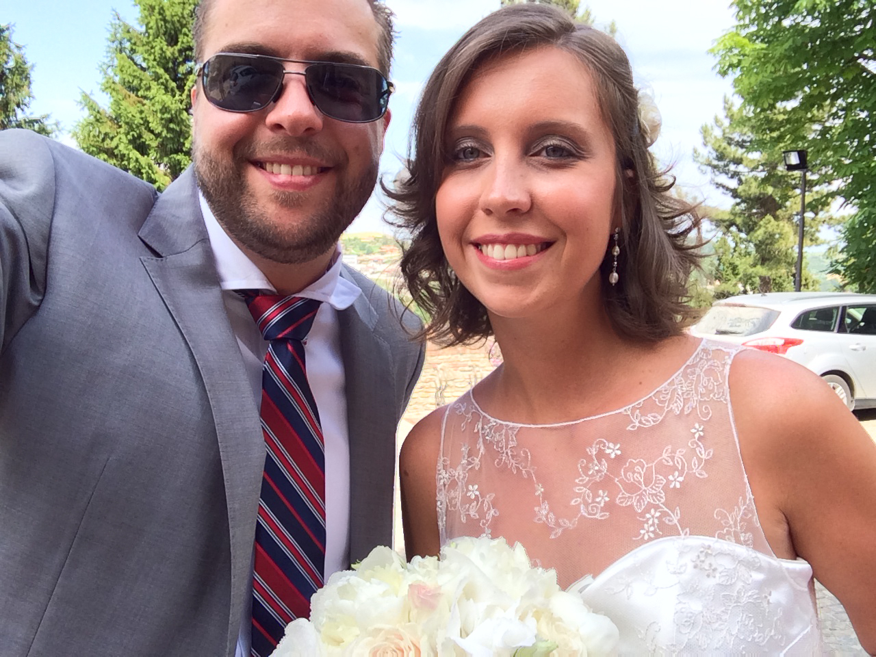 A smiling bride in a white dress holds a bouquet, posing for a selfie with a man in a suit.