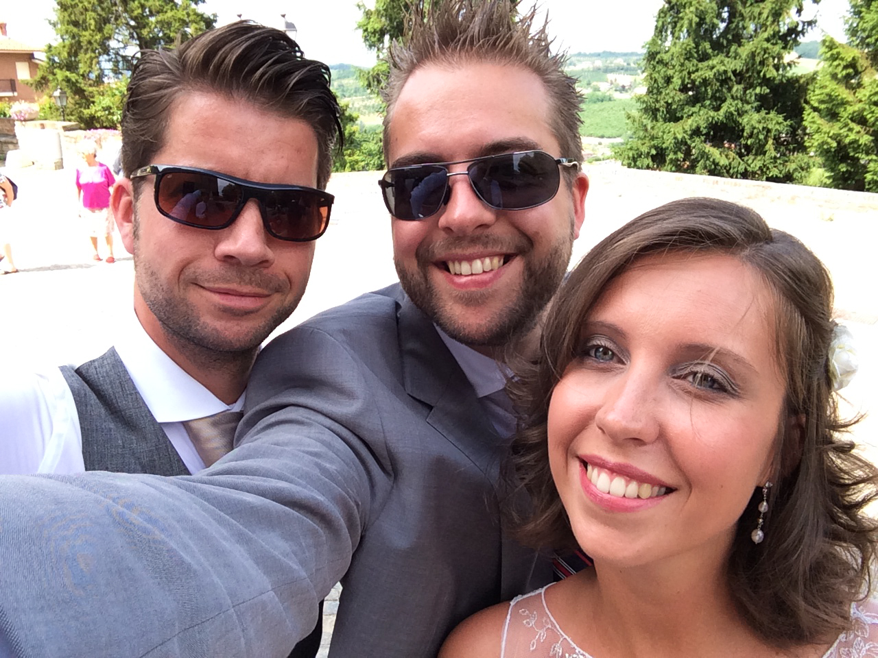 Three people dressed in wedding attire smile for a selfie outdoors, with the bride in the foreground.