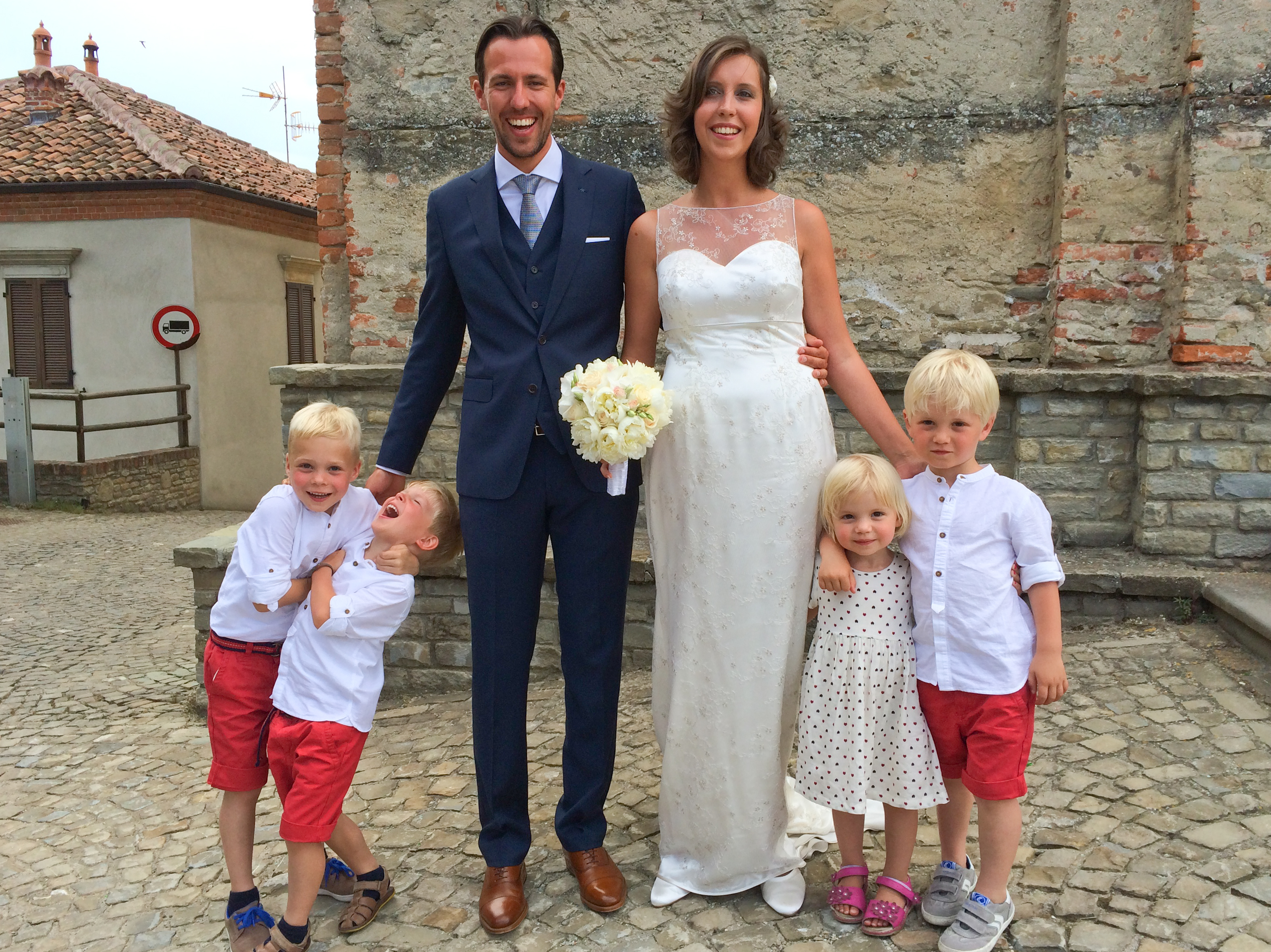 A bride and groom stand smiling with four young children, dressed in coordinated outfits, on a cobblestone street.