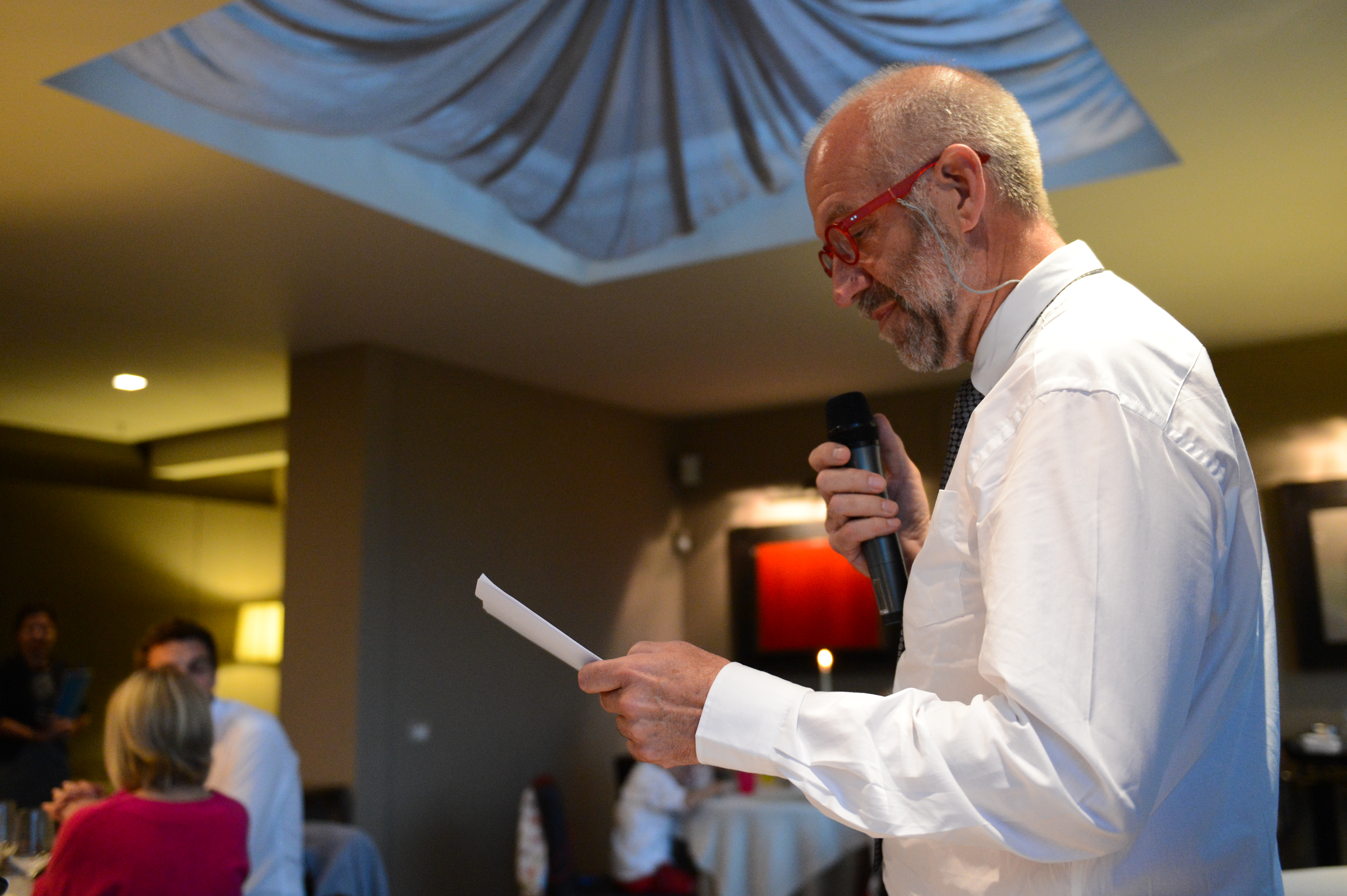 A man in a white shirt and red glasses gives a speech at a wedding, holding a microphone and paper.