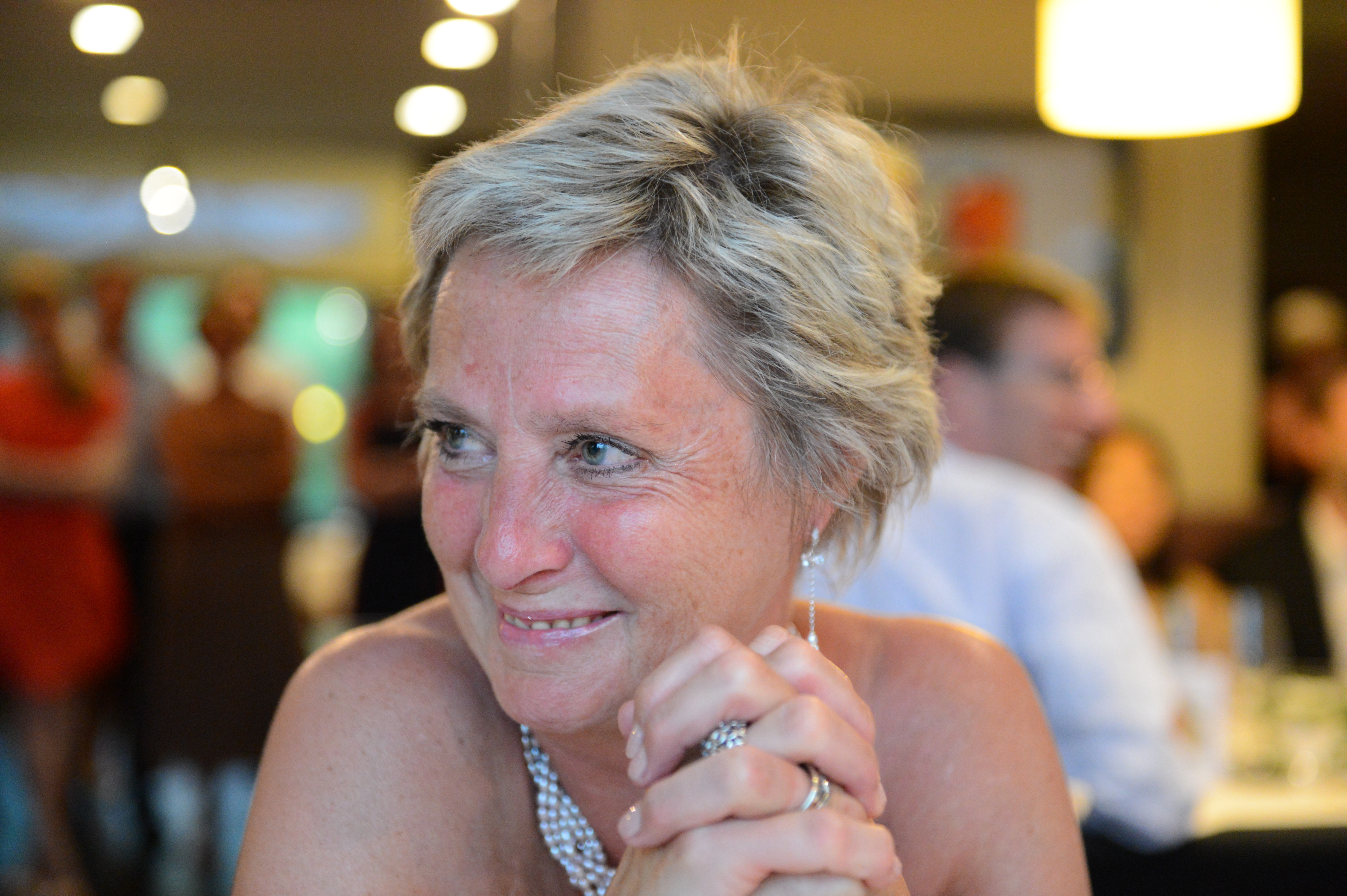 A woman with short blonde hair smiles and clasps her hands at a wedding reception.