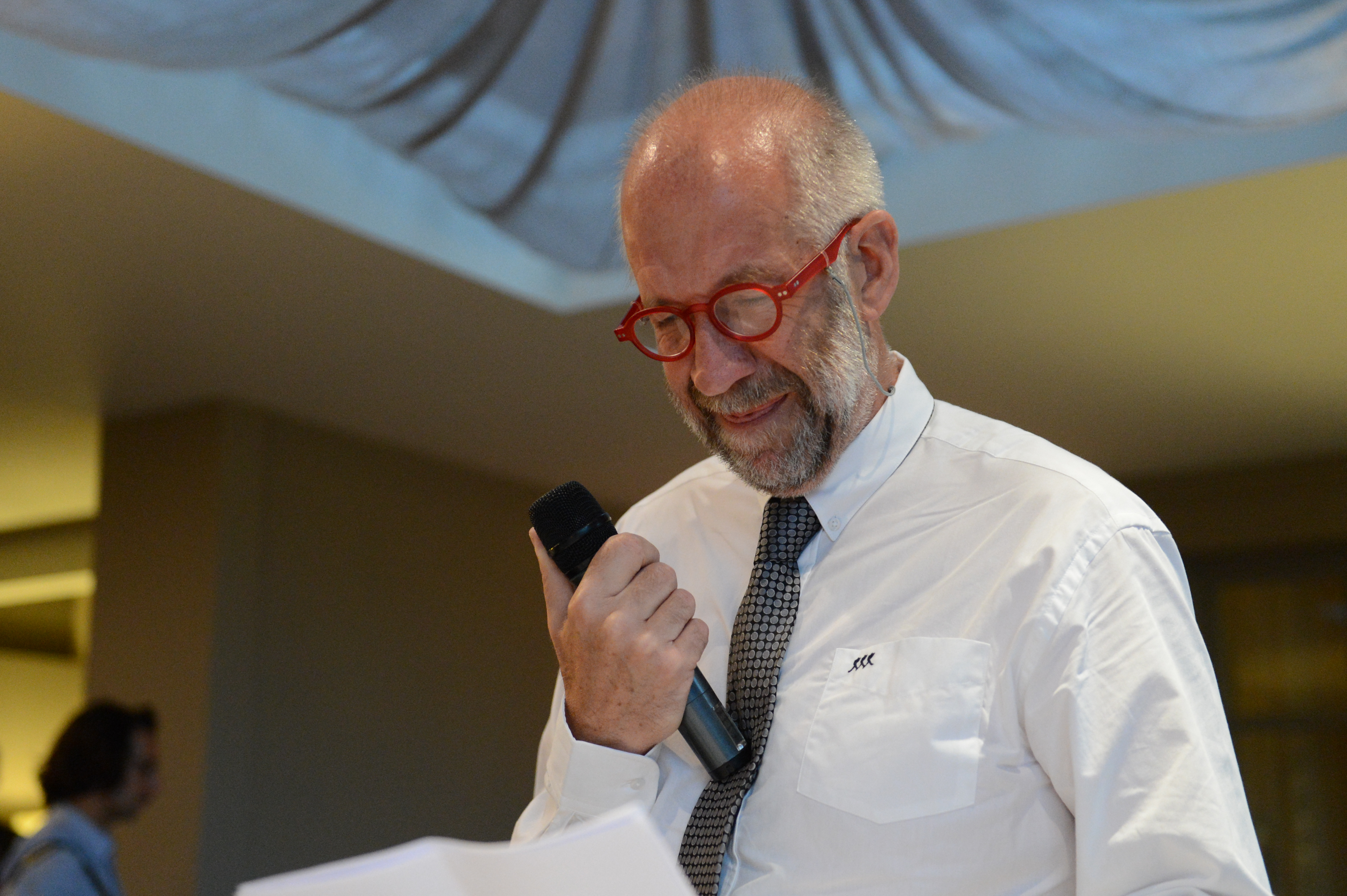 An older man in a white shirt and red glasses gives an emotional speech at a wedding, holding a microphone.