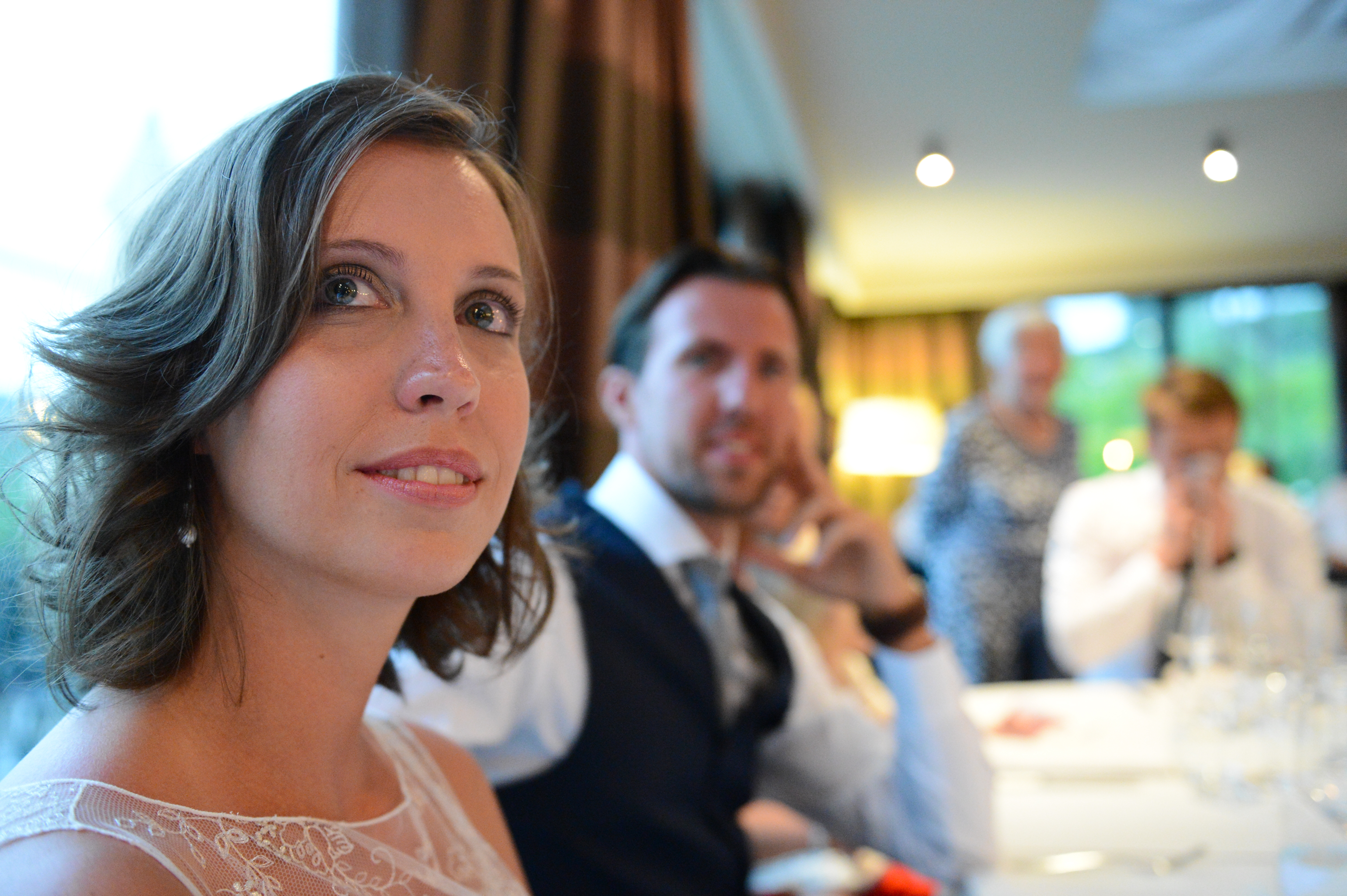 A woman in a wedding dress sits at a table, looking ahead, with guests in the background.