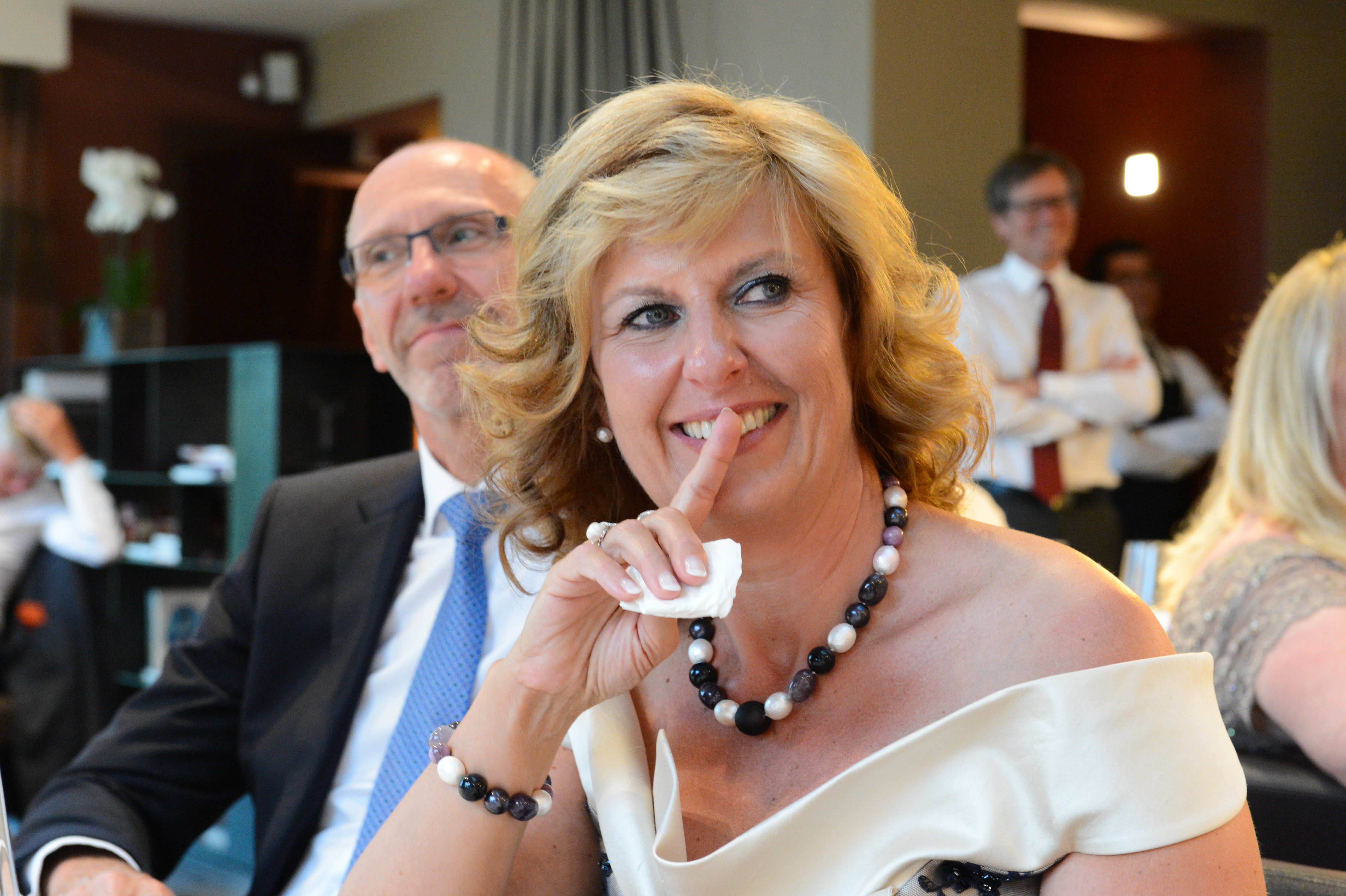 A woman in an elegant dress smiles while holding a tissue, with a man in a suit beside her.