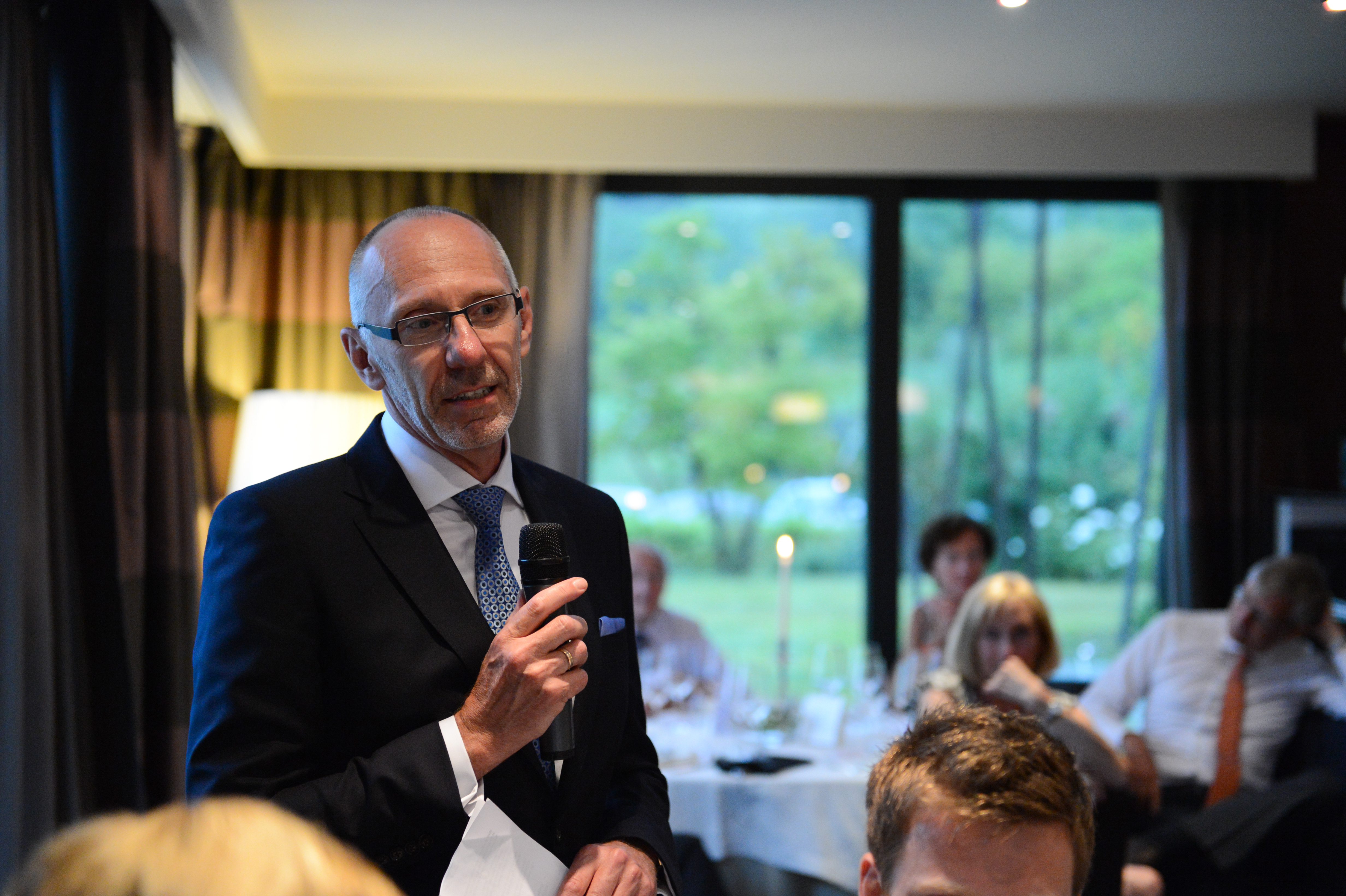 A man in a suit holds a microphone and gives a speech at a wedding reception.