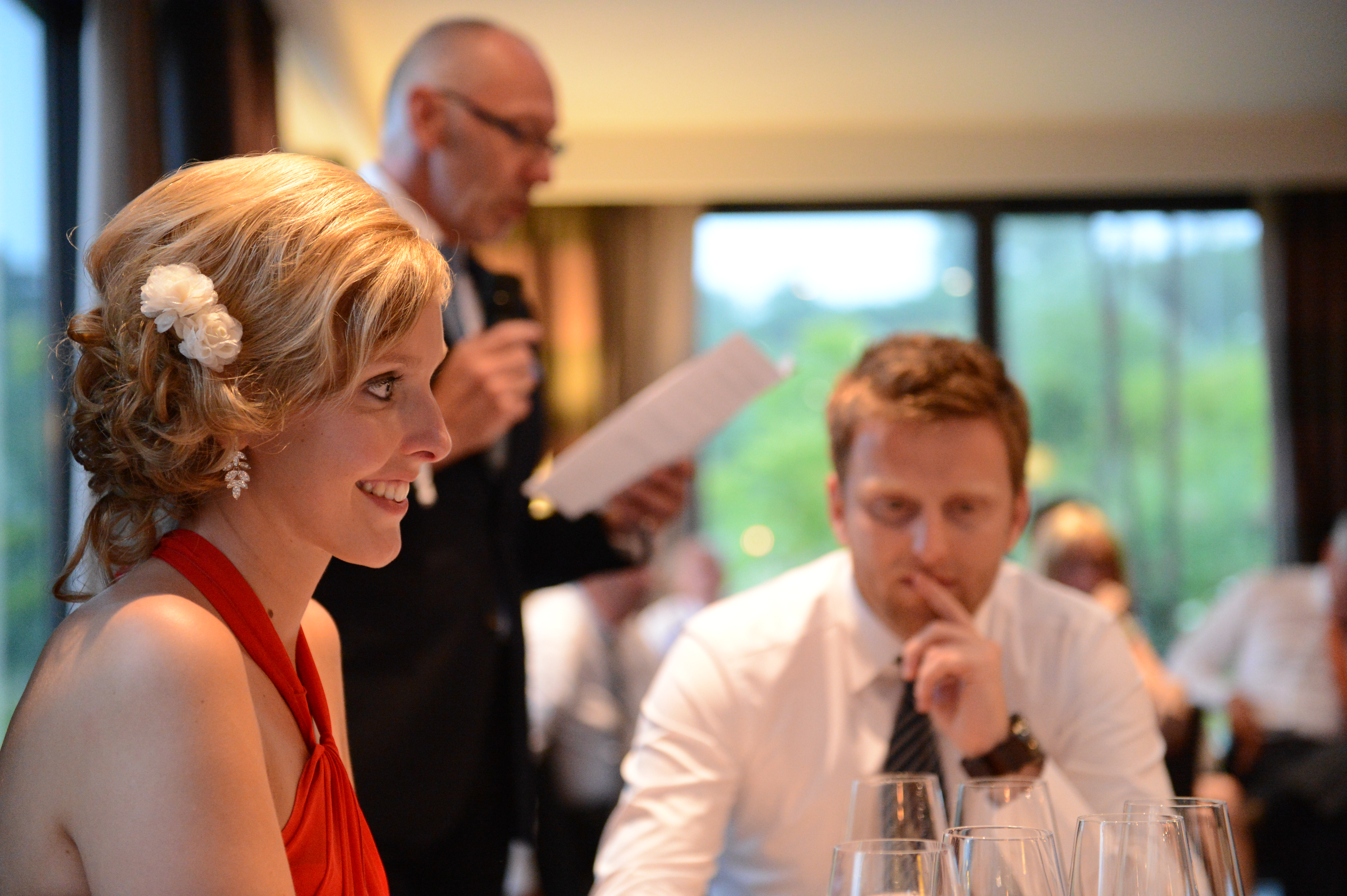 A woman in a red dress smiles while a man in a suit gives a speech at a wedding reception.