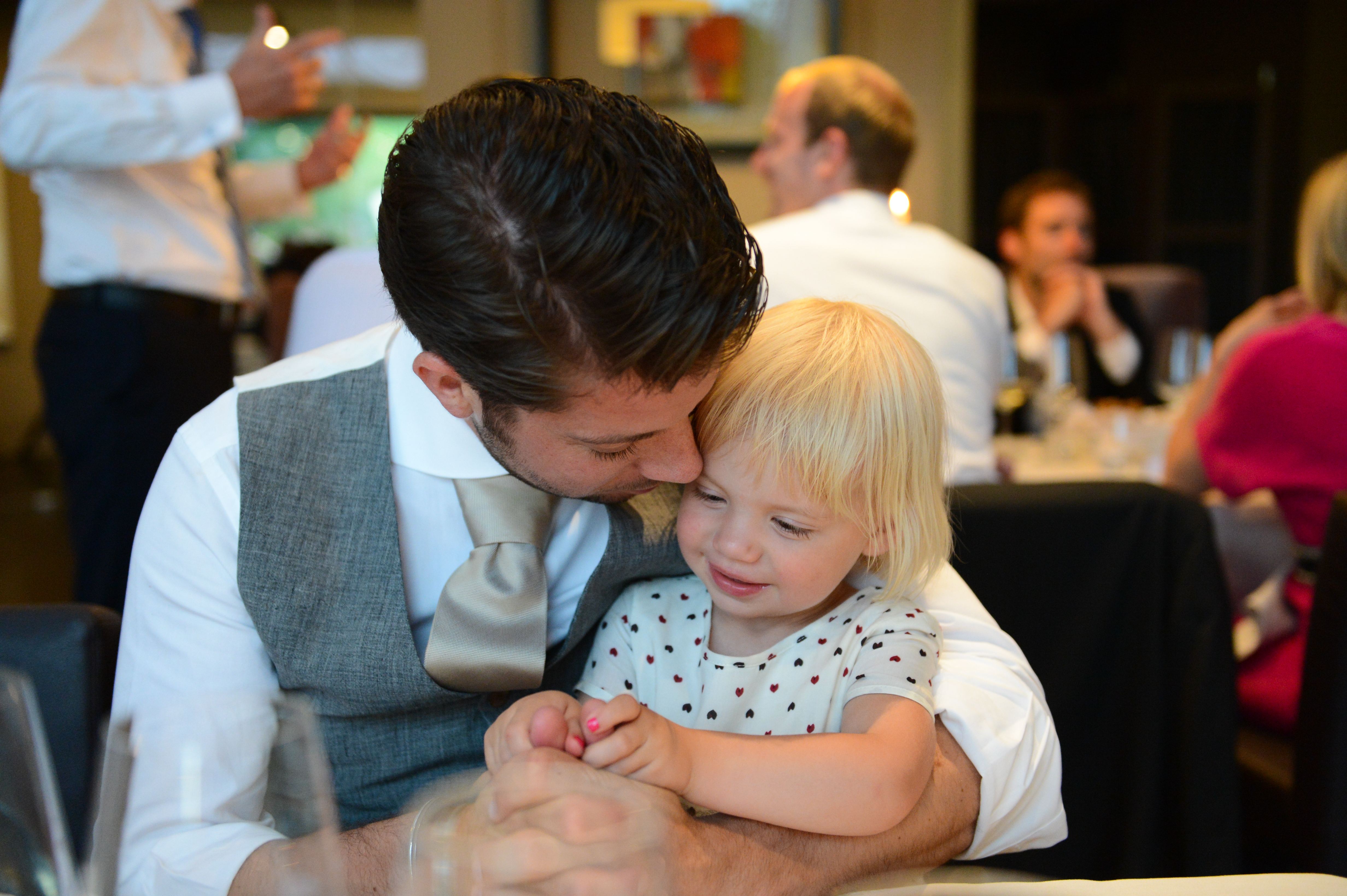 A man in a wedding vest hugs and kisses a smiling young child at a reception table.