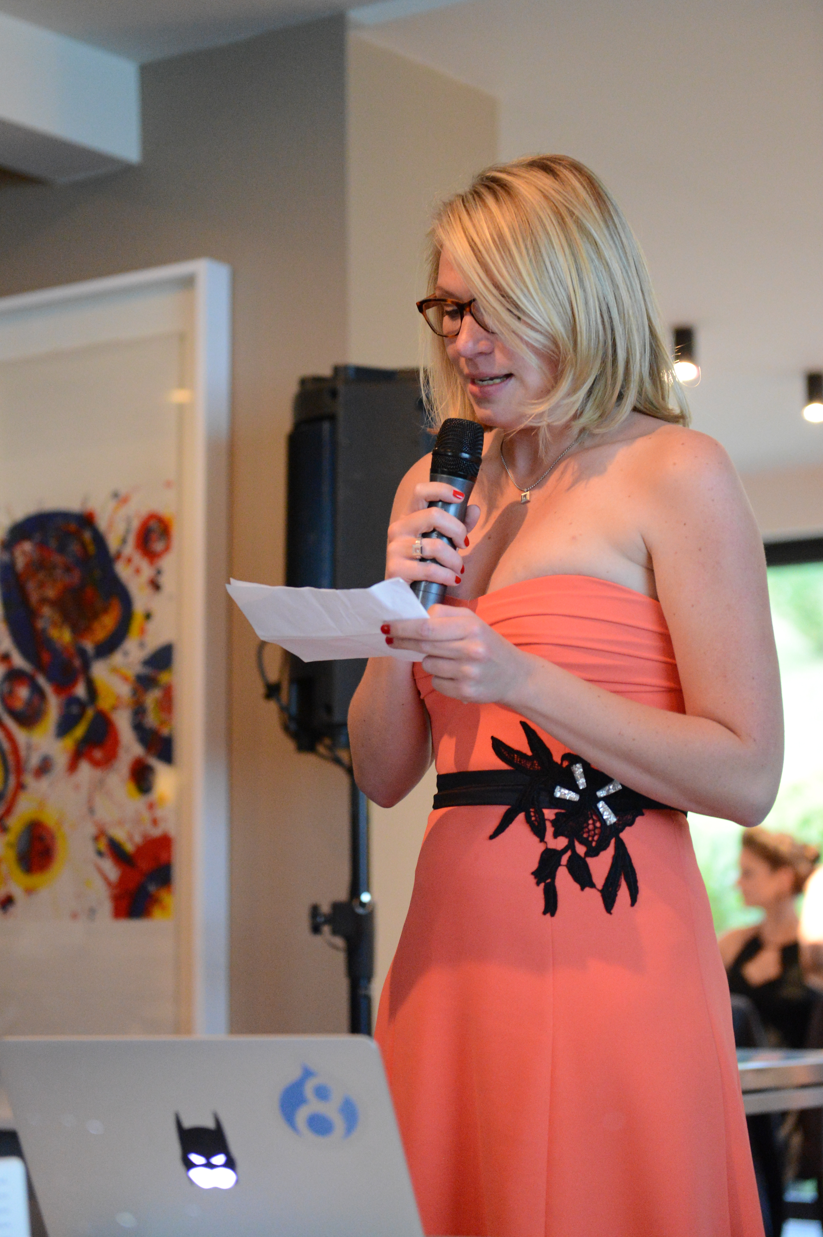 A woman in a coral dress holds a microphone and reads from a paper during a wedding speech.