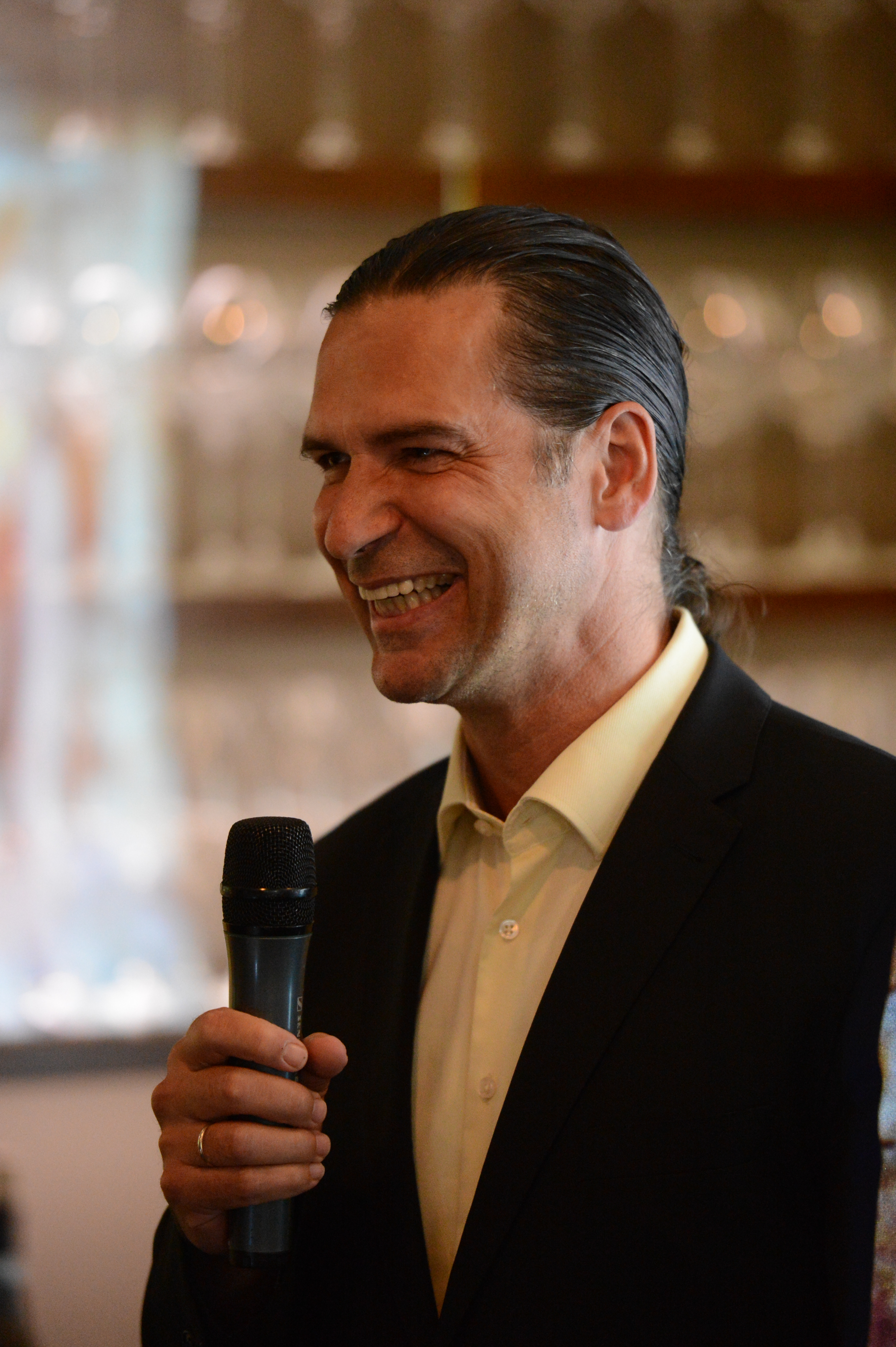 A man in a suit smiles while holding a microphone, speaking at a wedding reception.