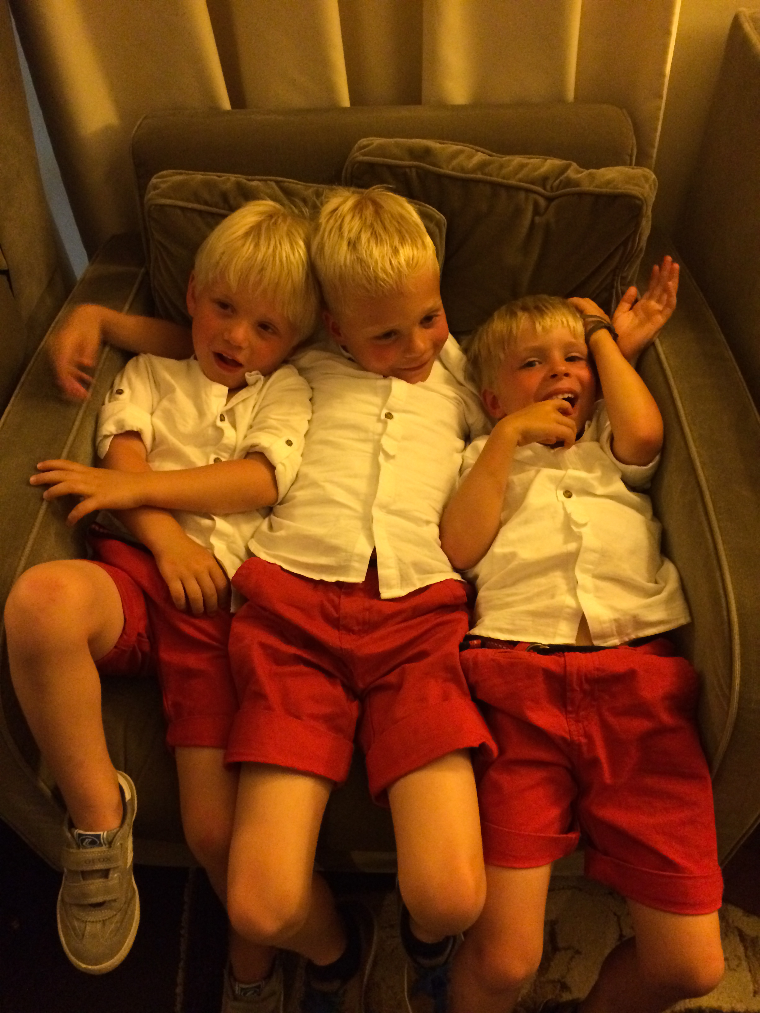 Three young boys in matching white shirts and red shorts sit closely together on a couch, smiling and laughing.