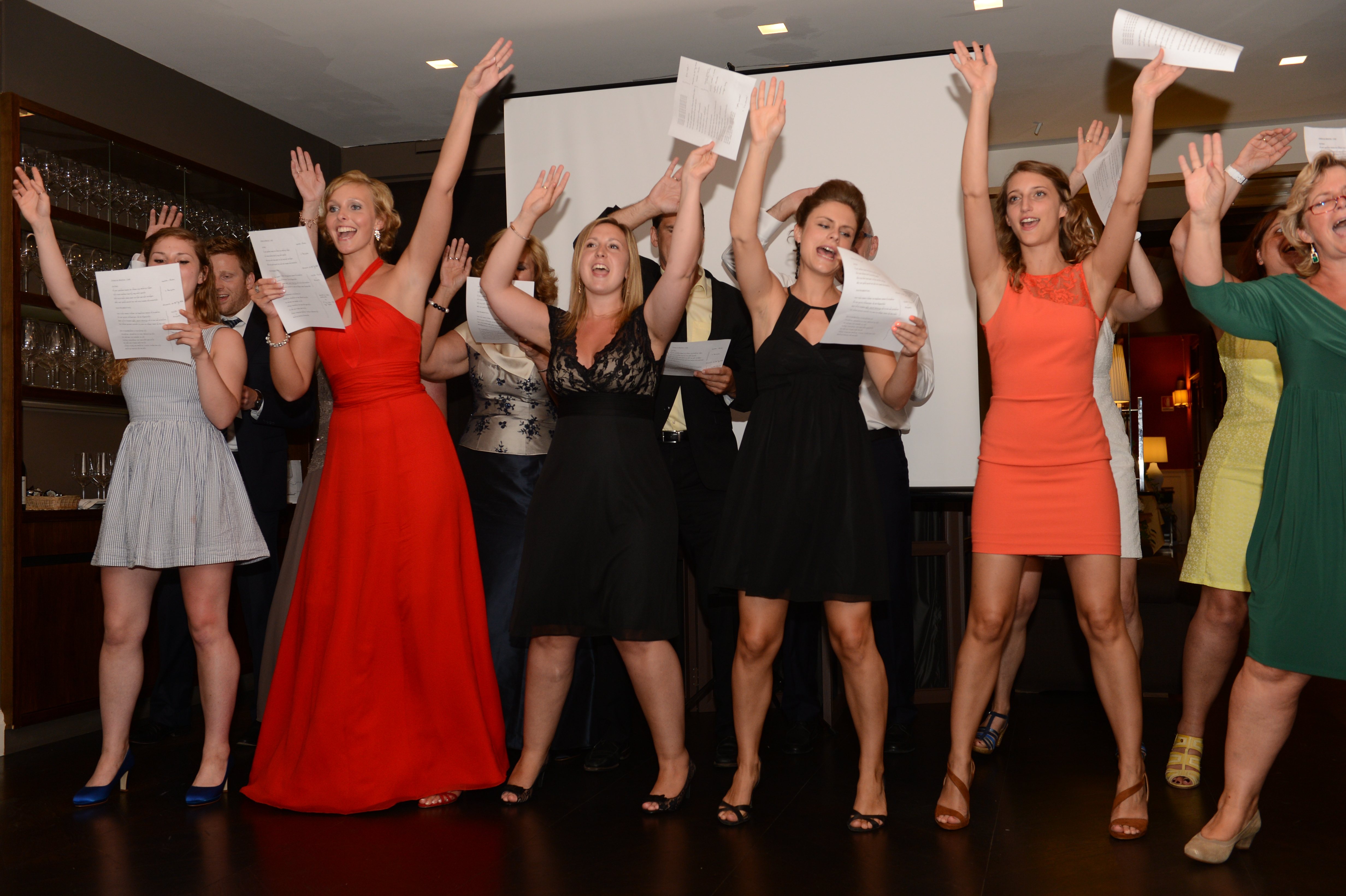 A group of wedding guests sings and raises their hands while holding papers during a celebration.