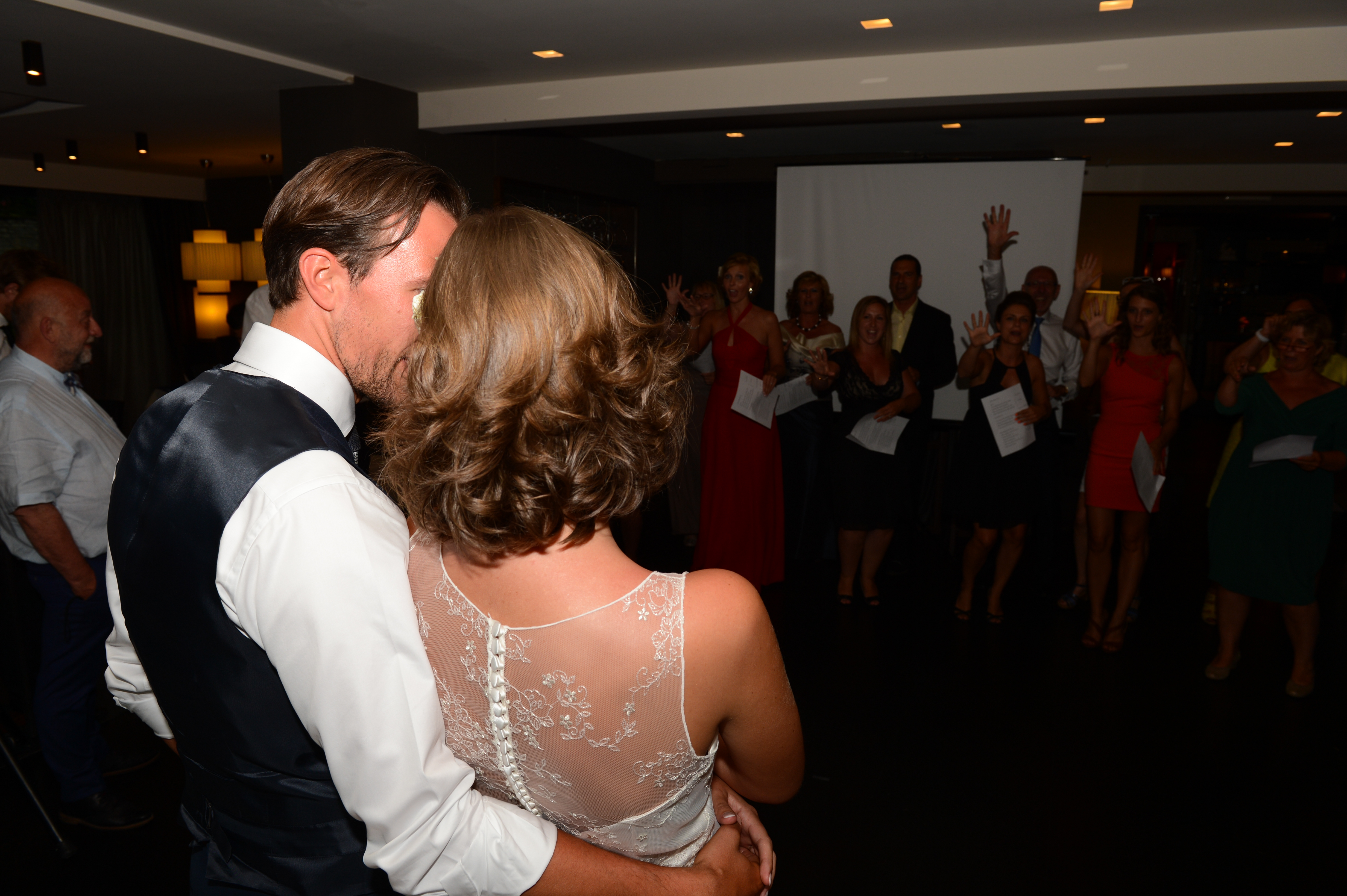 The bride and groom embrace while facing a group of wedding guests who are smiling and raising their hands.