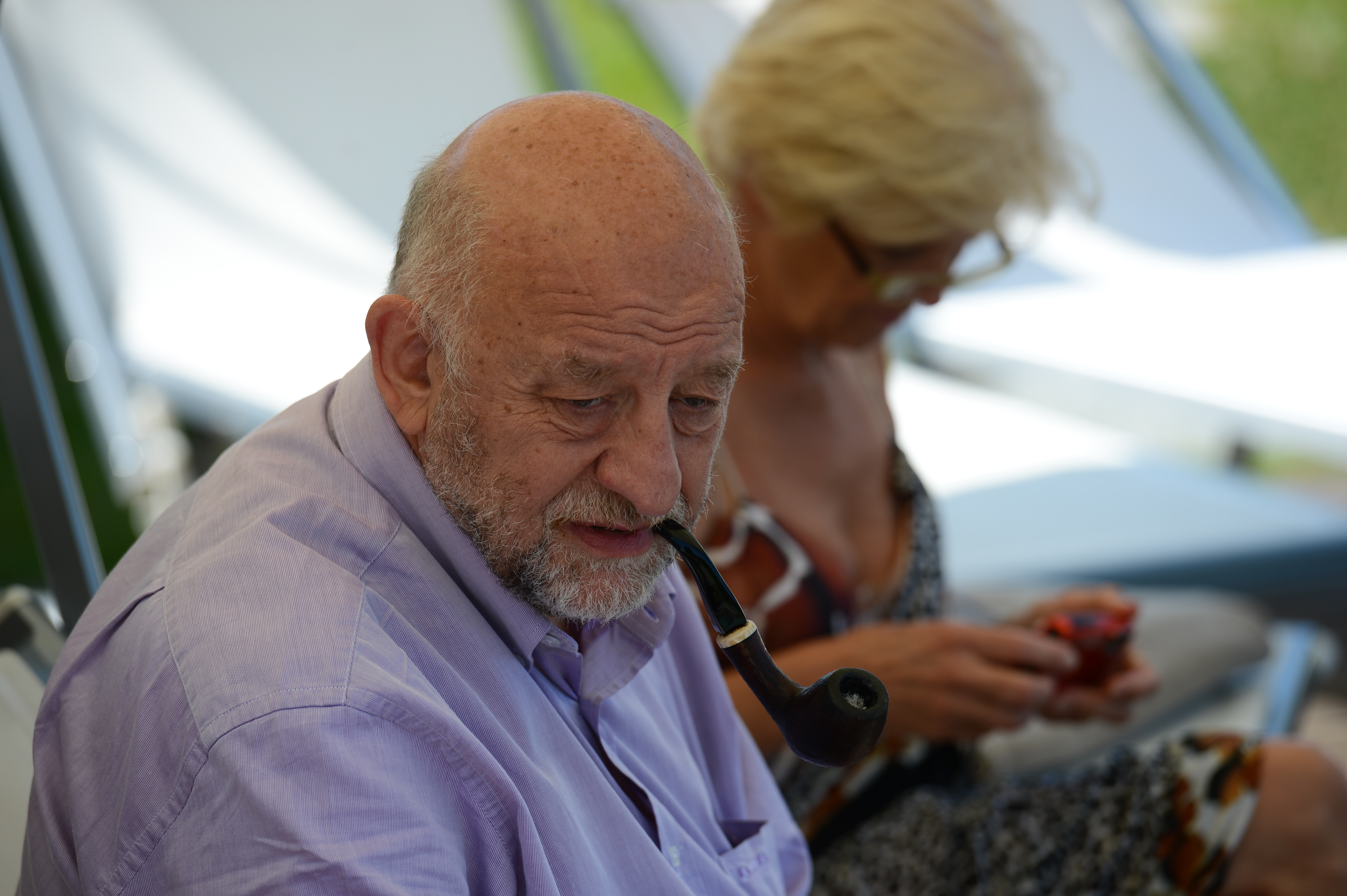 An older man in a light purple shirt smokes a pipe, while a woman behind him looks at her phone.