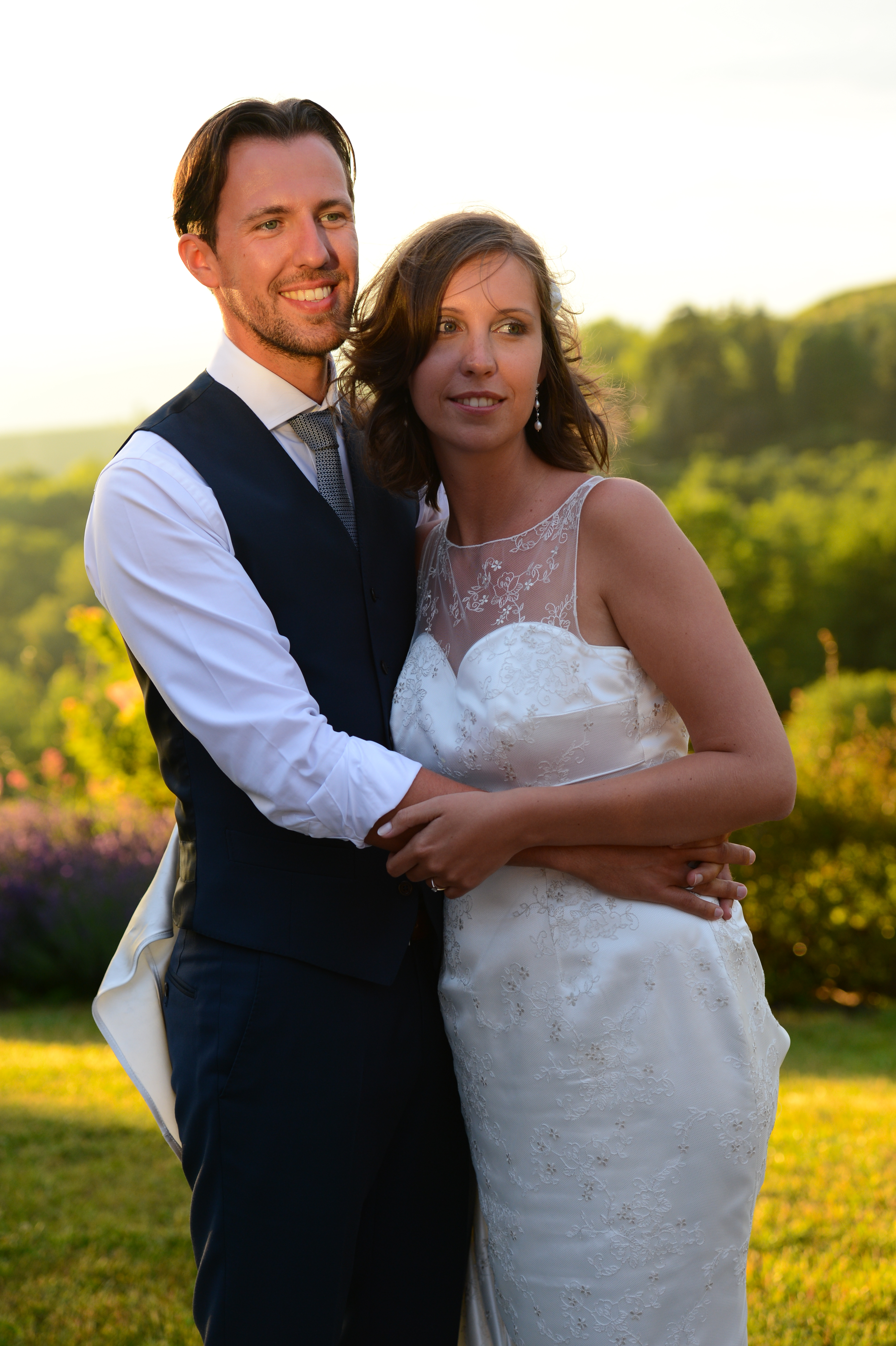 A bride and groom stand outdoors, embracing and smiling on their wedding day.