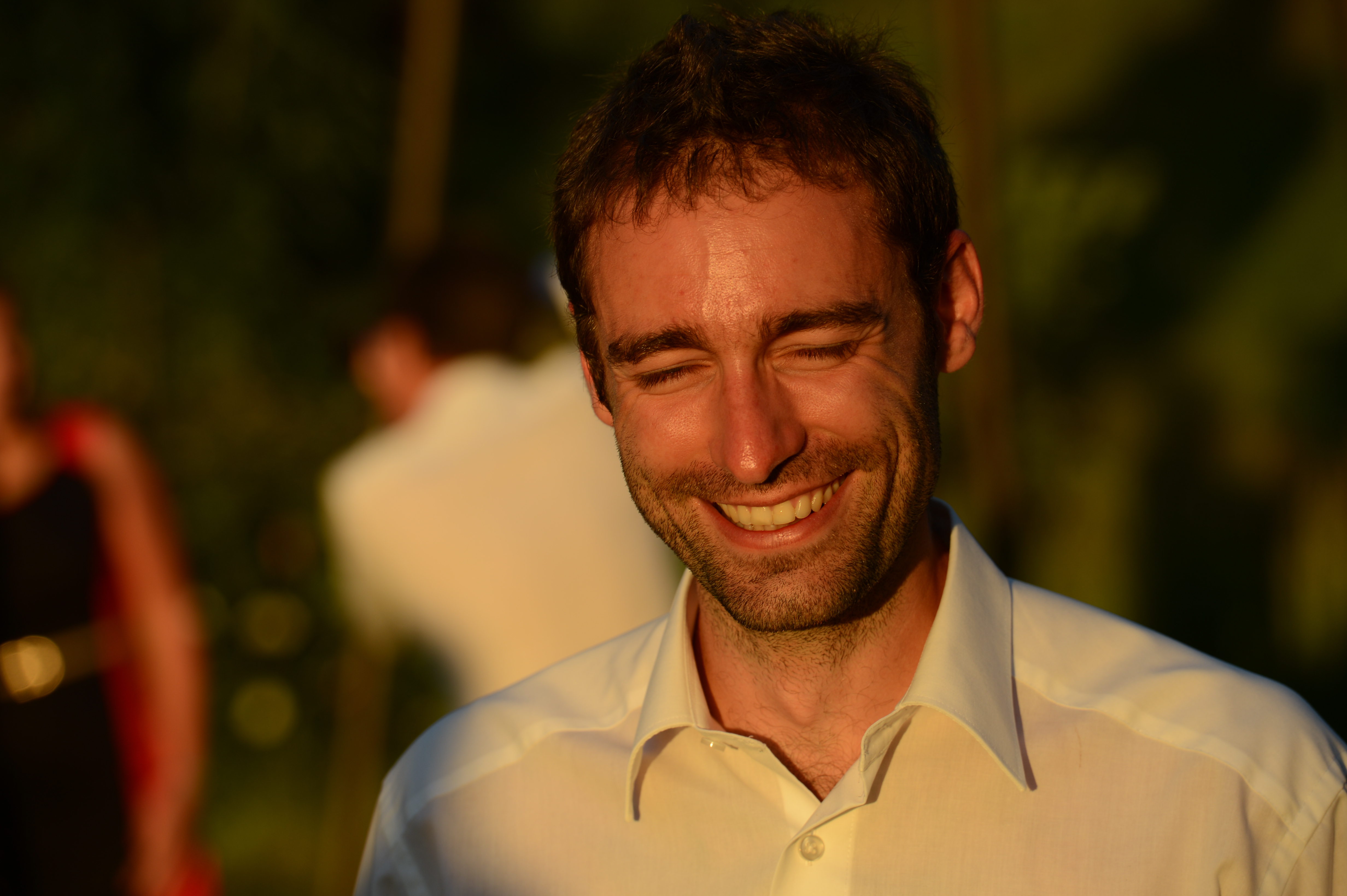 A man in a white shirt smiles with his eyes closed, standing outdoors in warm sunlight.