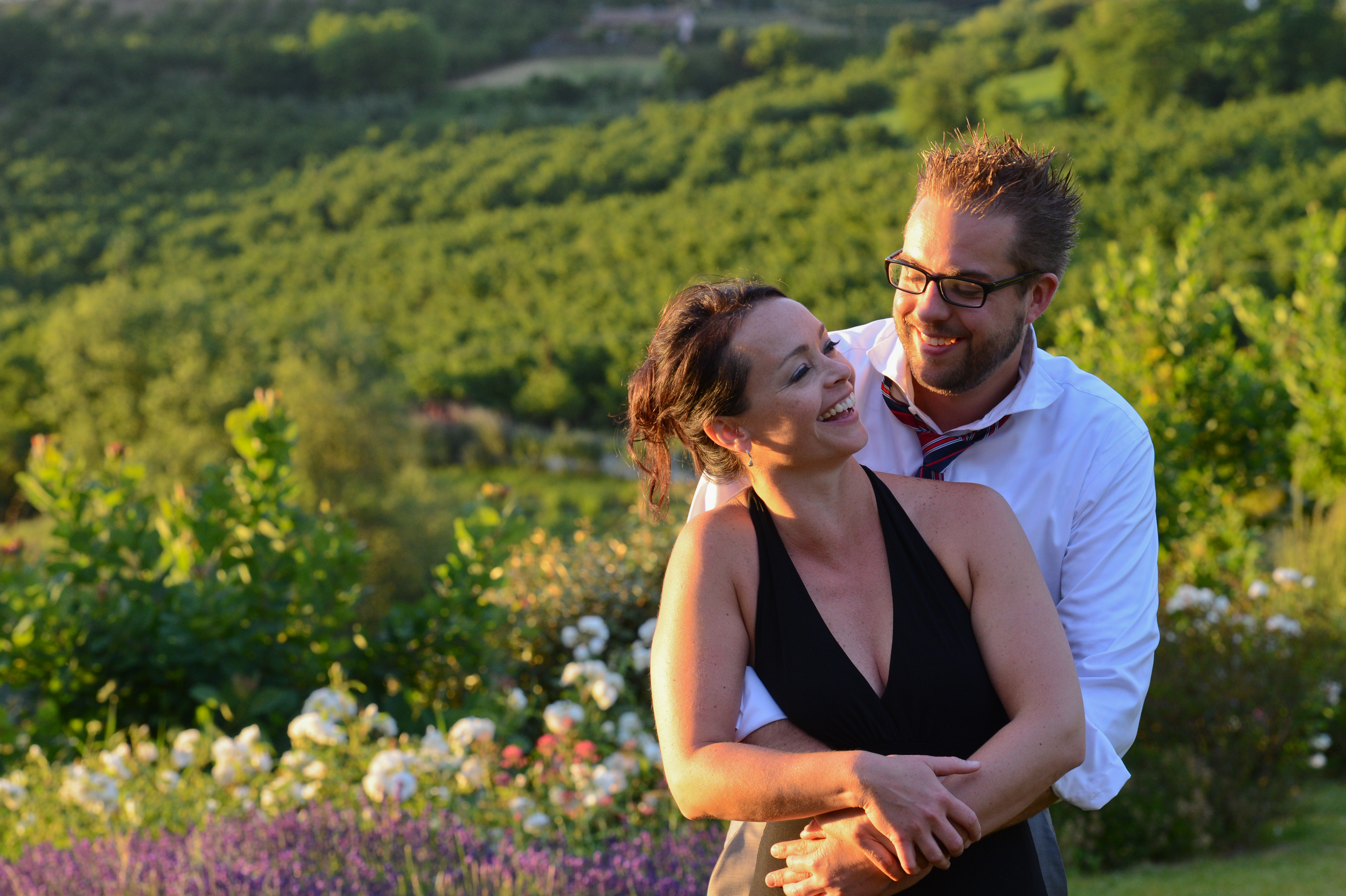 A smiling couple embraces outdoors, with the man holding the woman from behind during a wedding celebration.