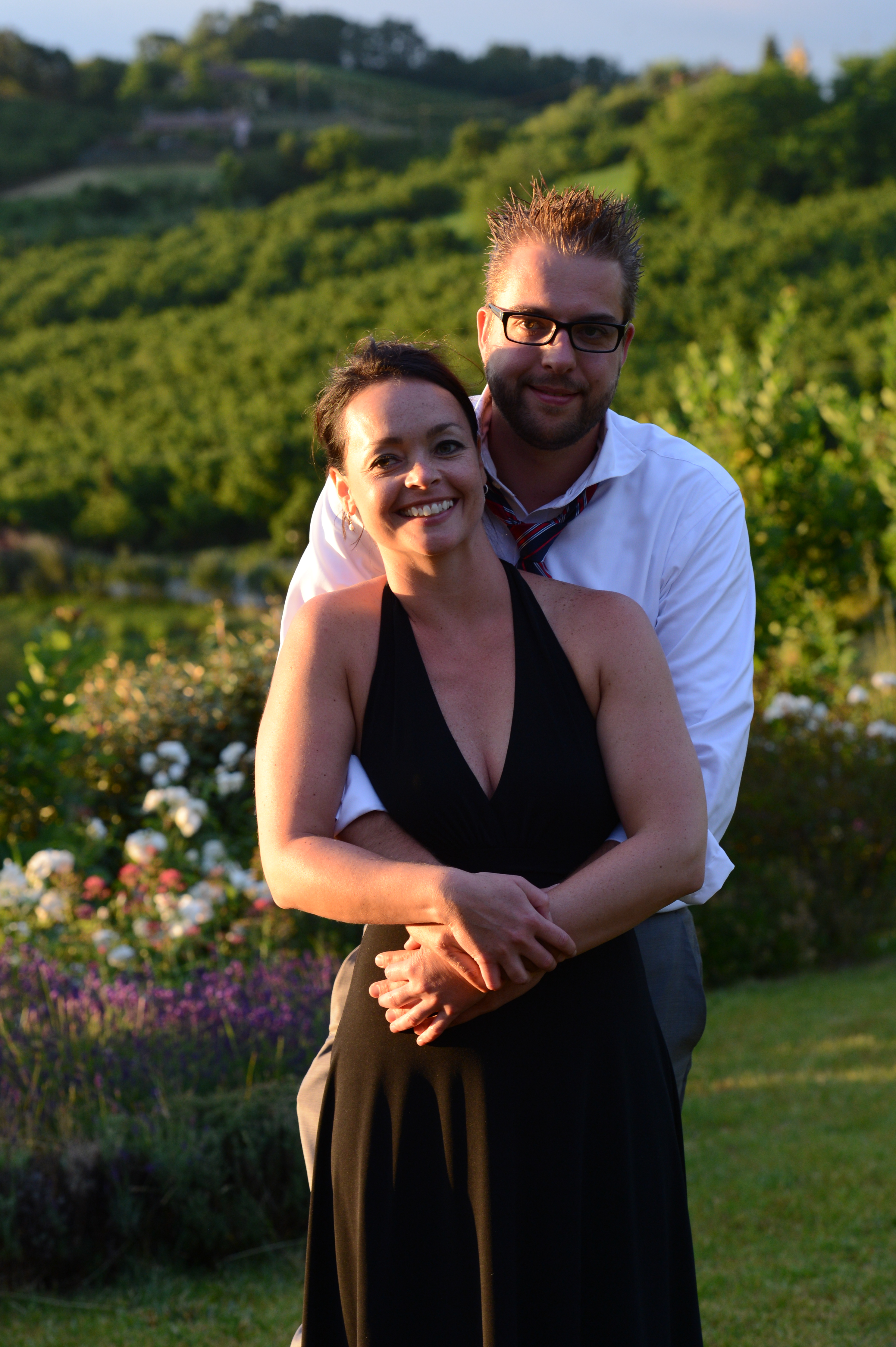 A smiling couple poses outdoors, with the man standing behind the woman and wrapping his arms around her.