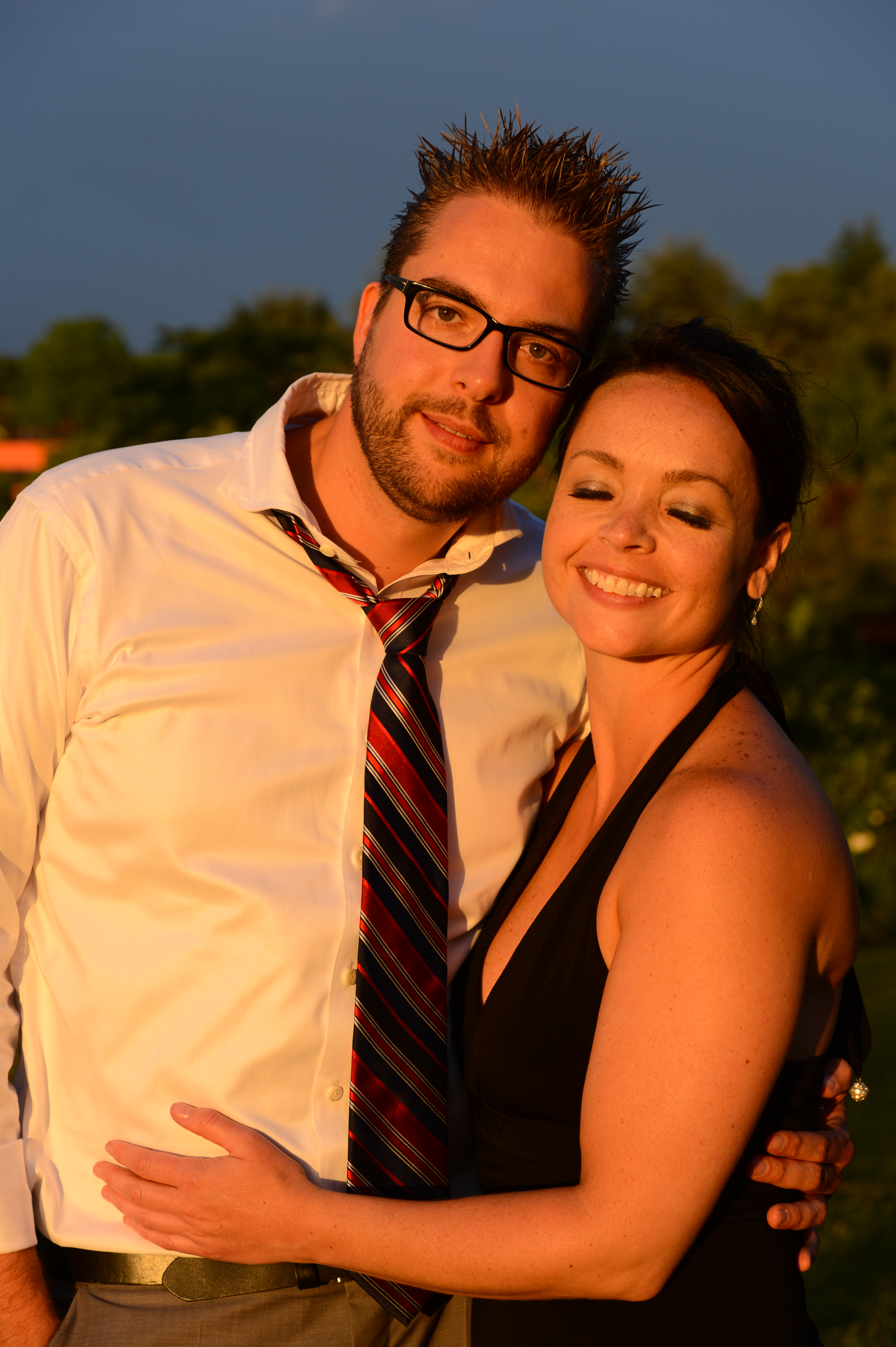 A couple embraces and smiles at the camera during a wedding celebration outdoors.
