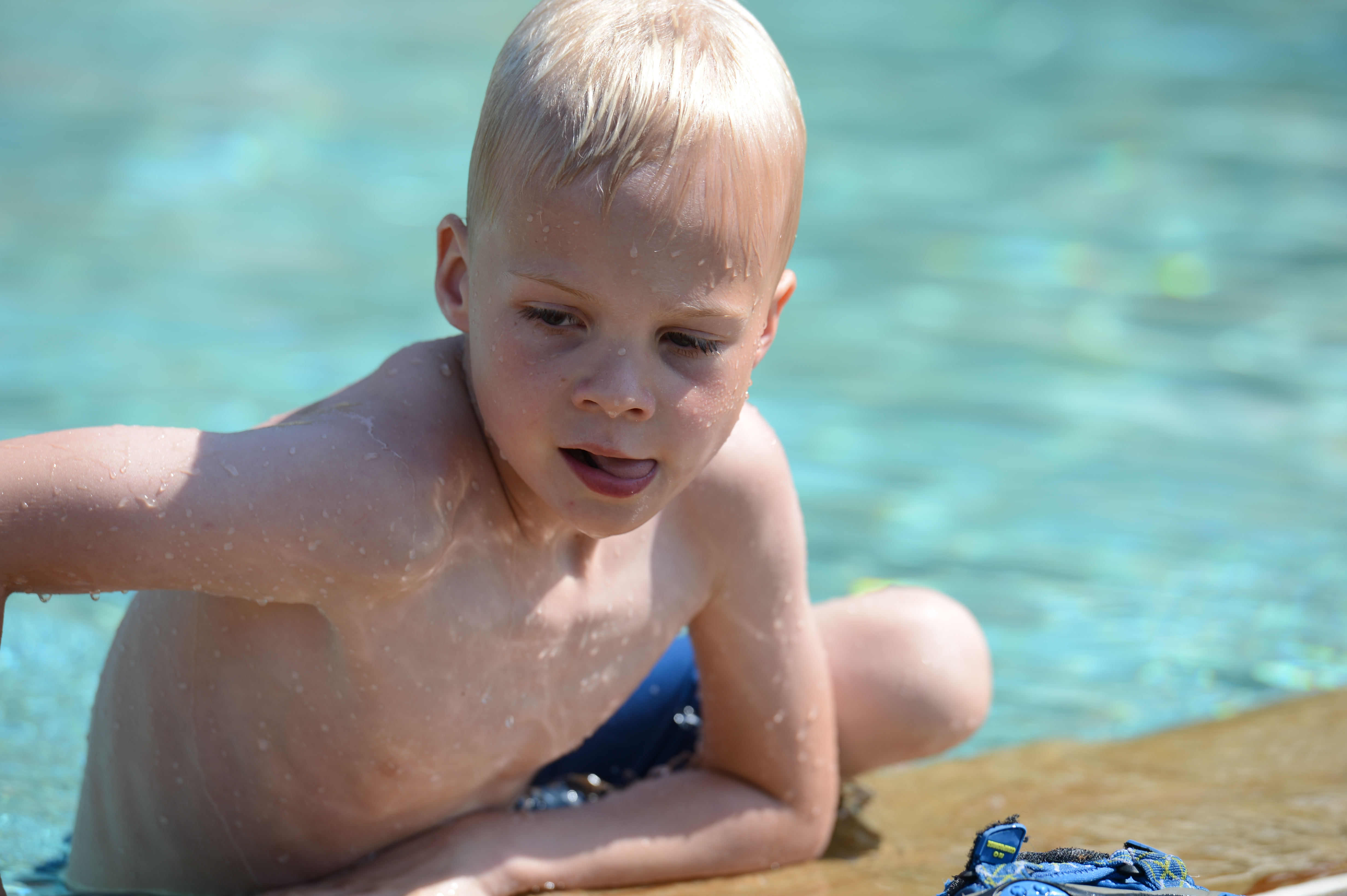 A young boy with wet skin sits on the edge of a pool, wearing blue swim trunks.