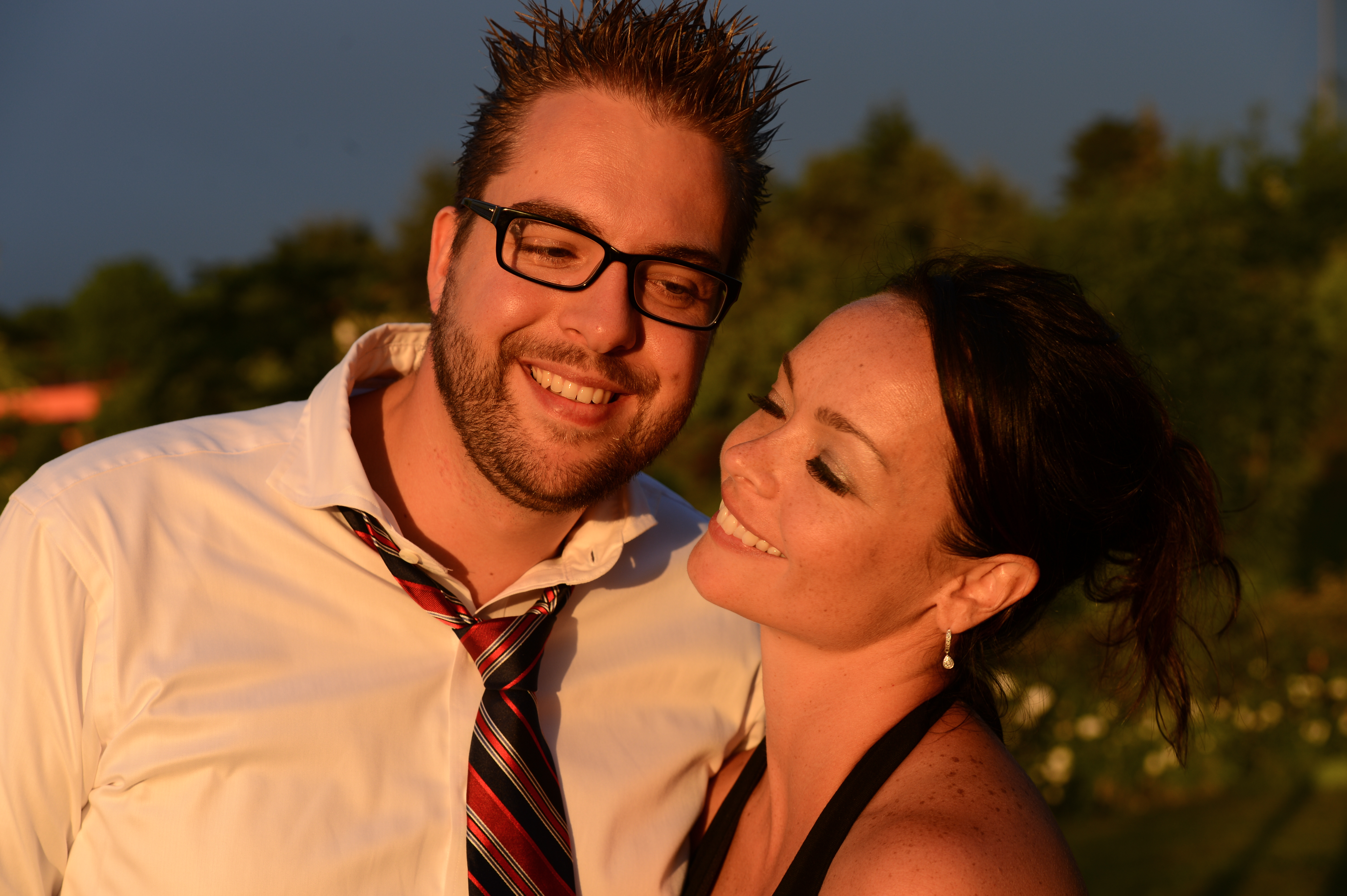 A smiling man in a loosened tie and glasses stands beside a woman looking at him with a happy expression.