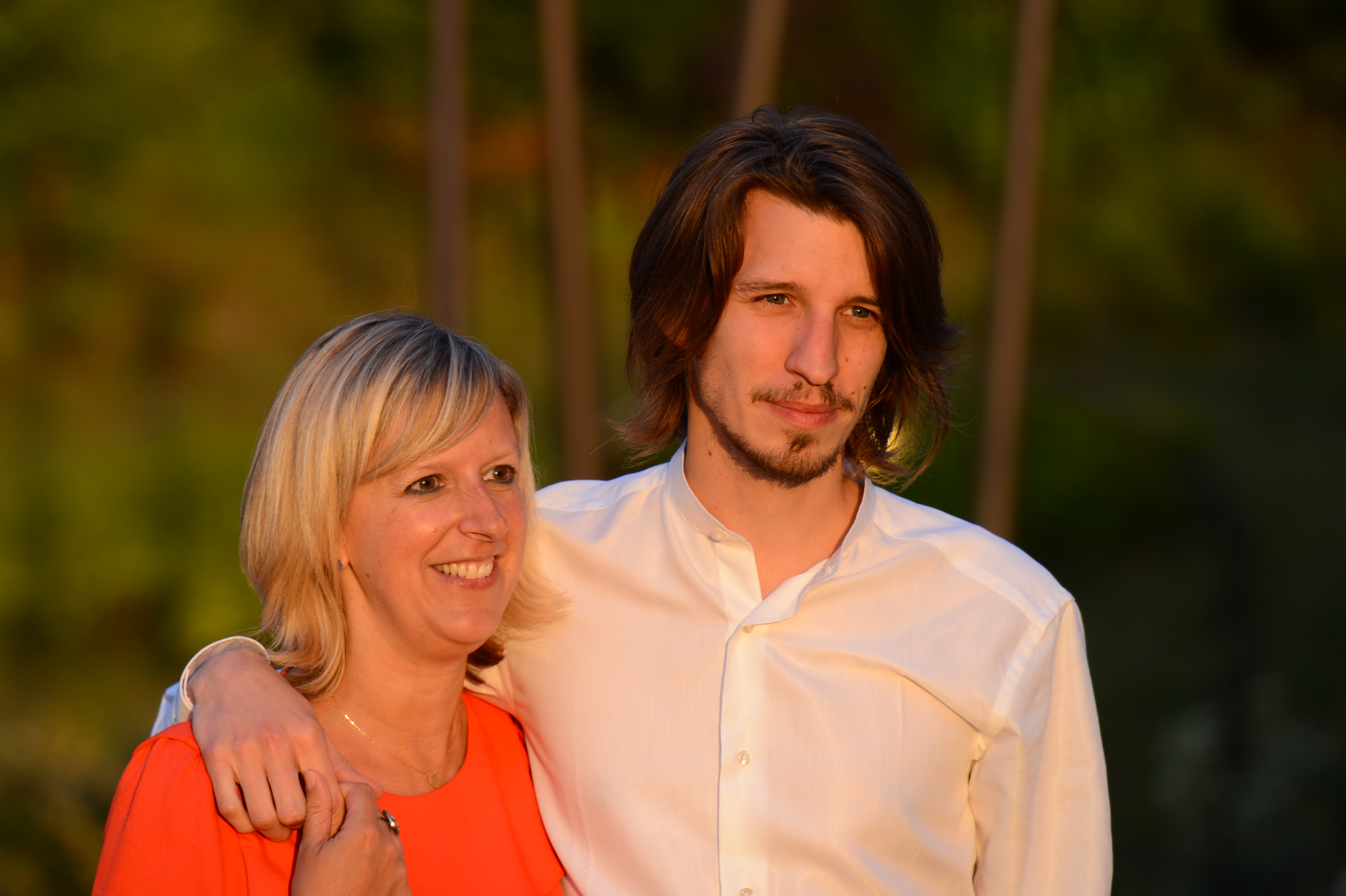 A man in a white shirt puts his arm around a smiling woman in an orange dress at a wedding.