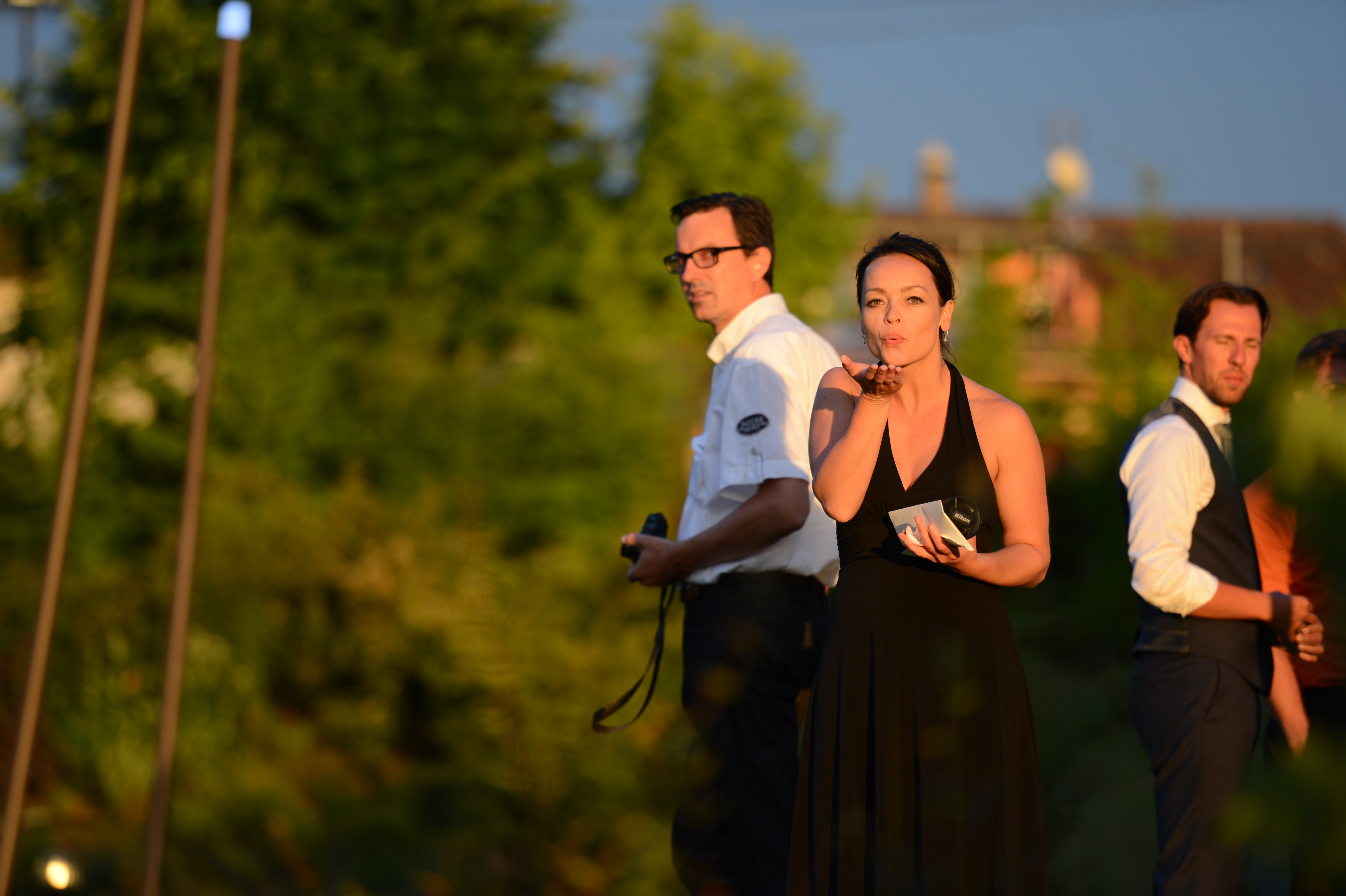 A woman in a black dress blows a kiss while holding small cards at an outdoor wedding event.