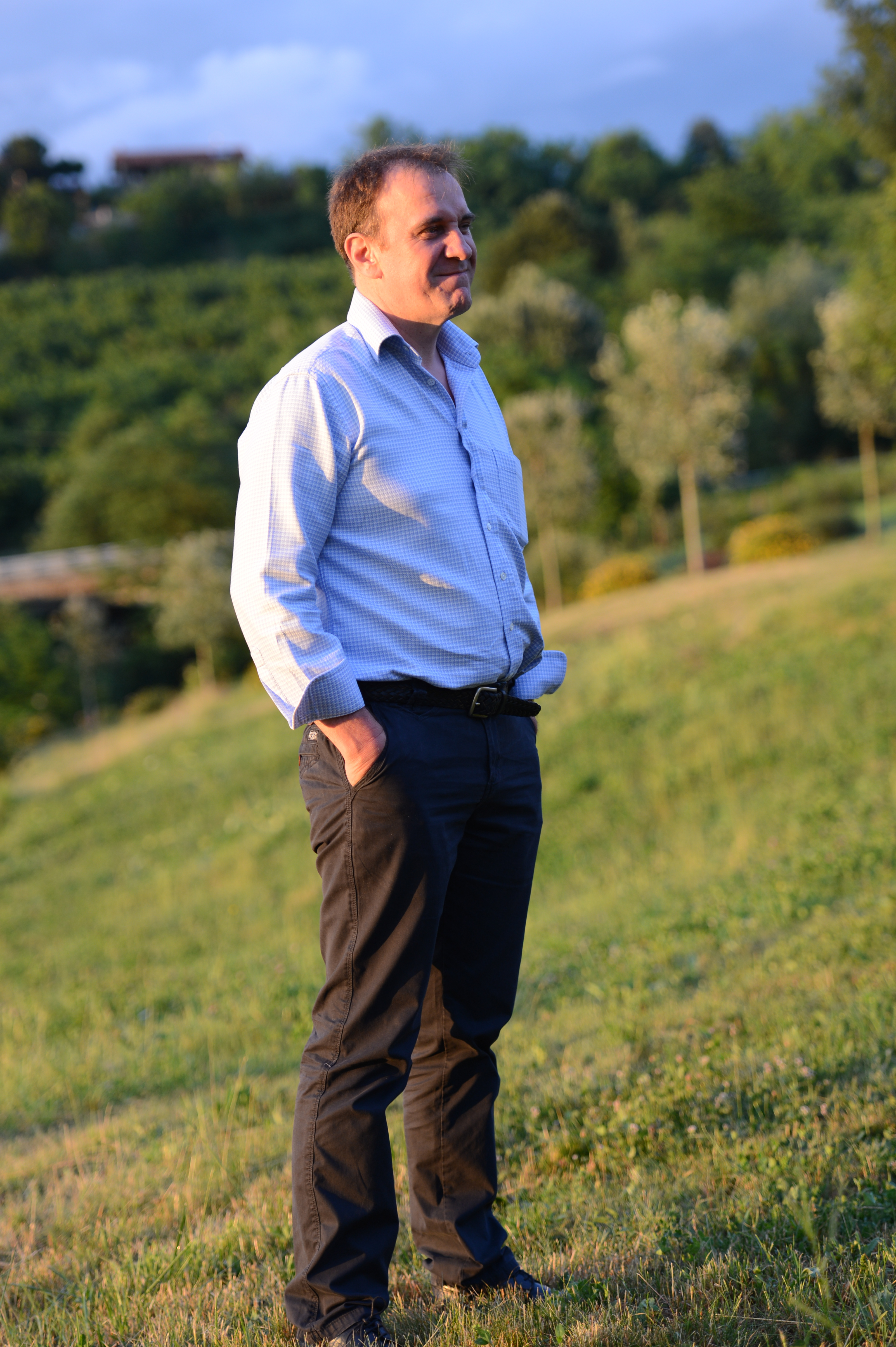 A man in a dress shirt and dark pants stands on a grassy hill, smiling and looking off-camera.
