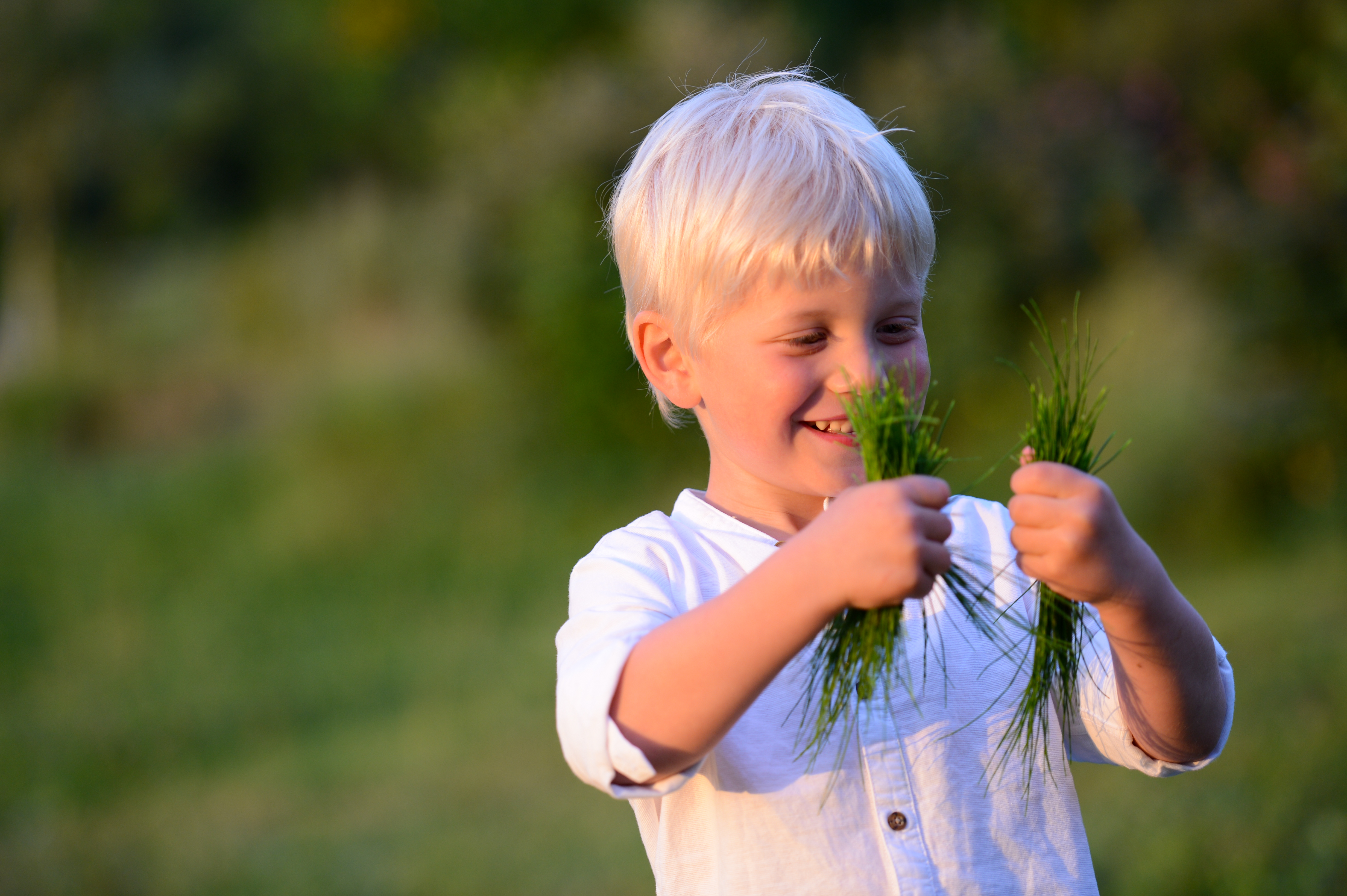 A young boy in a white shirt smiles while holding handfuls of grass at an outdoor wedding.