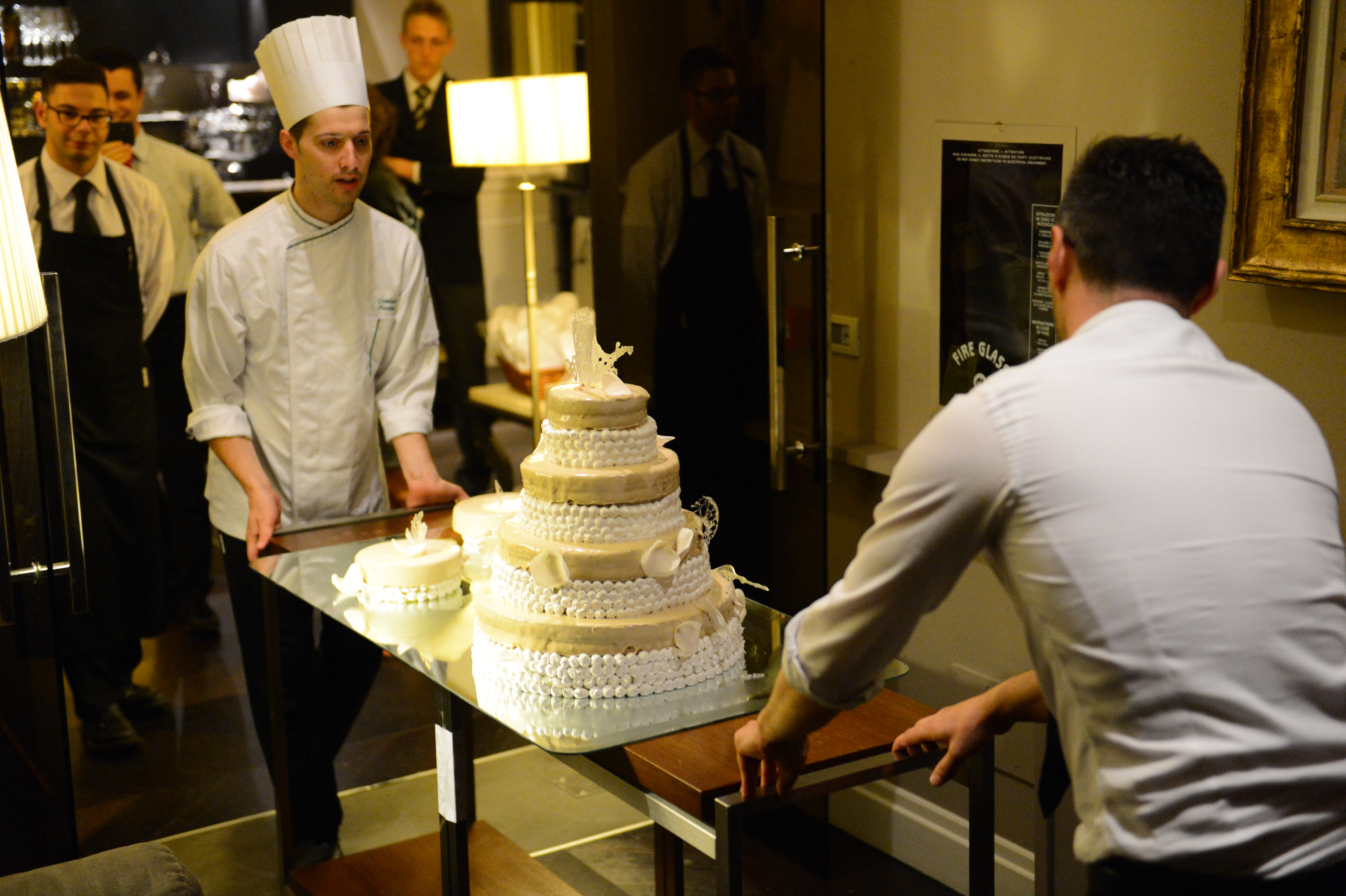 Two men carefully transport a large, decorated wedding cake on a glass table, with staff watching in the background.