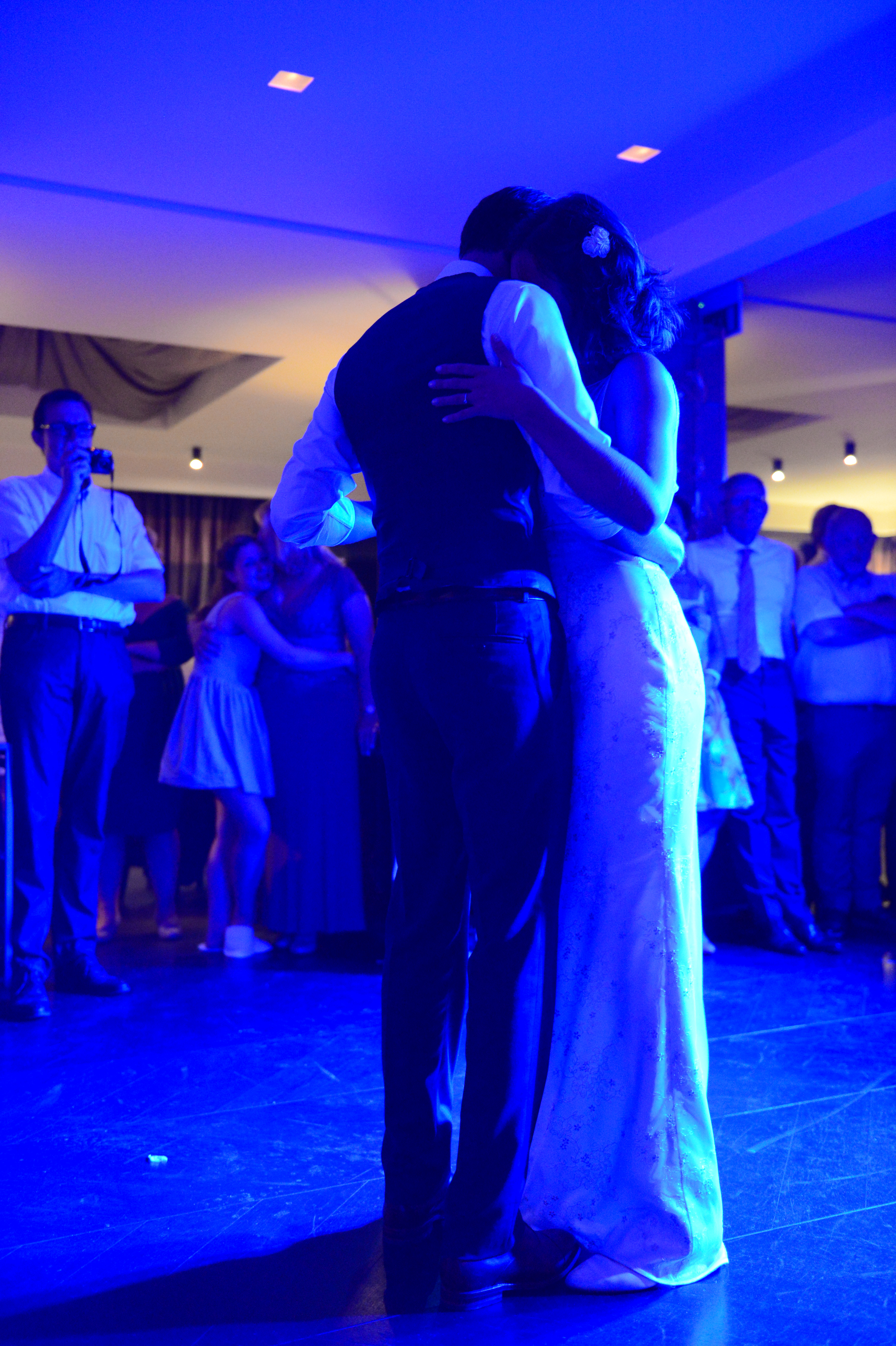 The bride and groom share a close embrace while dancing, surrounded by wedding guests watching.