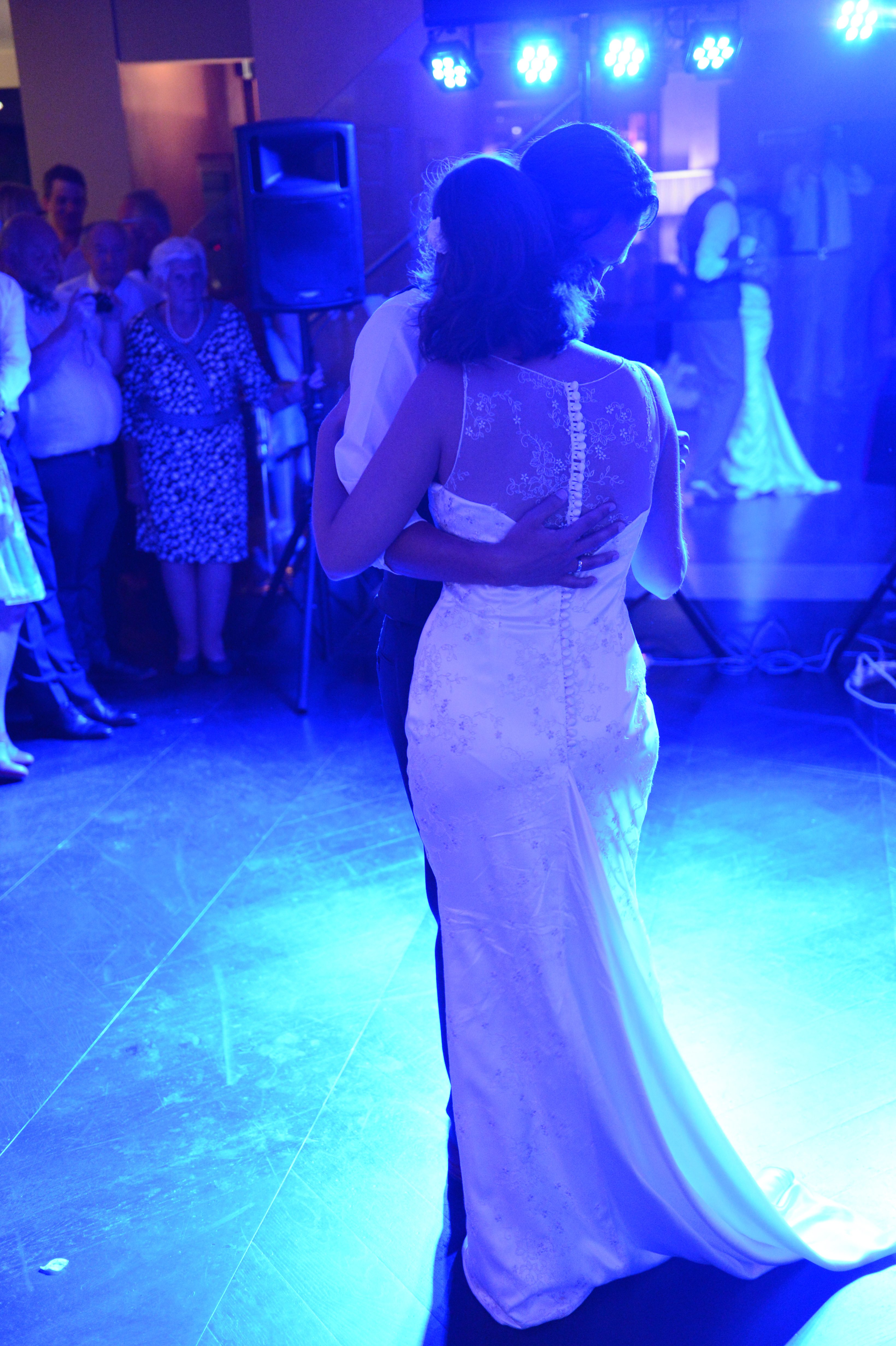 The bride and groom share their first dance, embracing on the dance floor under blue lighting, with guests watching.